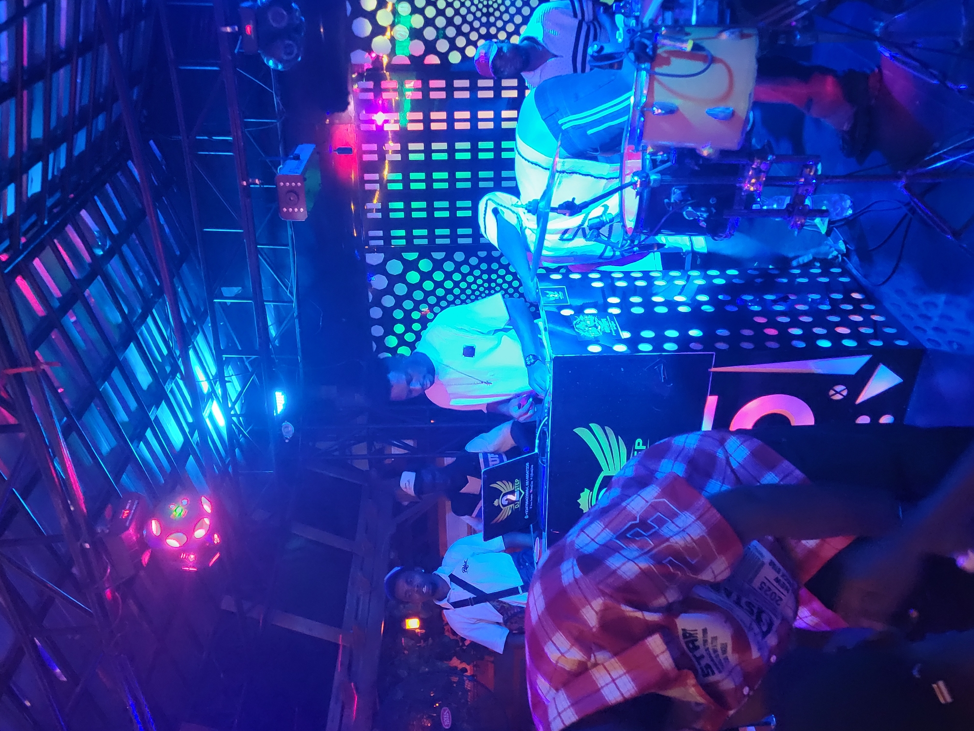 Inside what appears to be a nightclub or entertainment venue in Obosi, Nigeria, several individuals are visible under vibrant, colorful lighting. A man in a red plaid shirt stands in the foreground, partially obscuring the view. Further back, two men are positioned at a DJ setup with illuminated panels and equipment. Above them, a man sits atop a structure or platform. The environment is decorated with neon lights and disco balls, indicating a lively atmosphere.