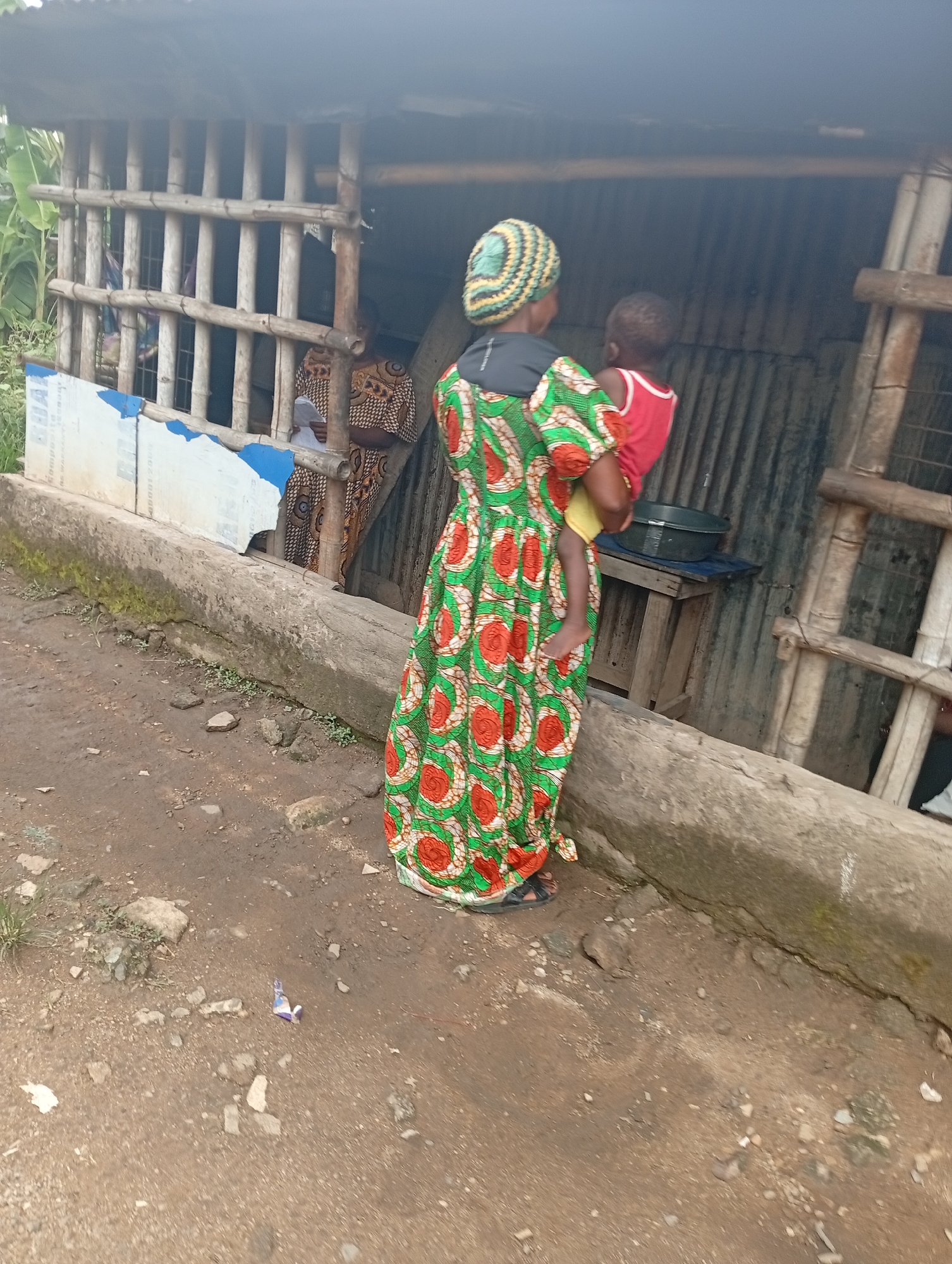 In Owerri, Nigeria, a woman in a colorful green and red patterned dress and a headwrap is holding a child in a red shirt and yellow shorts. They are standing next to a weathered concrete structure. Behind them is a makeshift shelter or building with bamboo framing and corrugated metal walls. Inside, another woman in a patterned dress is visible, seemingly holding a document. A ladder leans against the building. A table with a dark-colored bowl sits inside. The ground is dirt and stone.