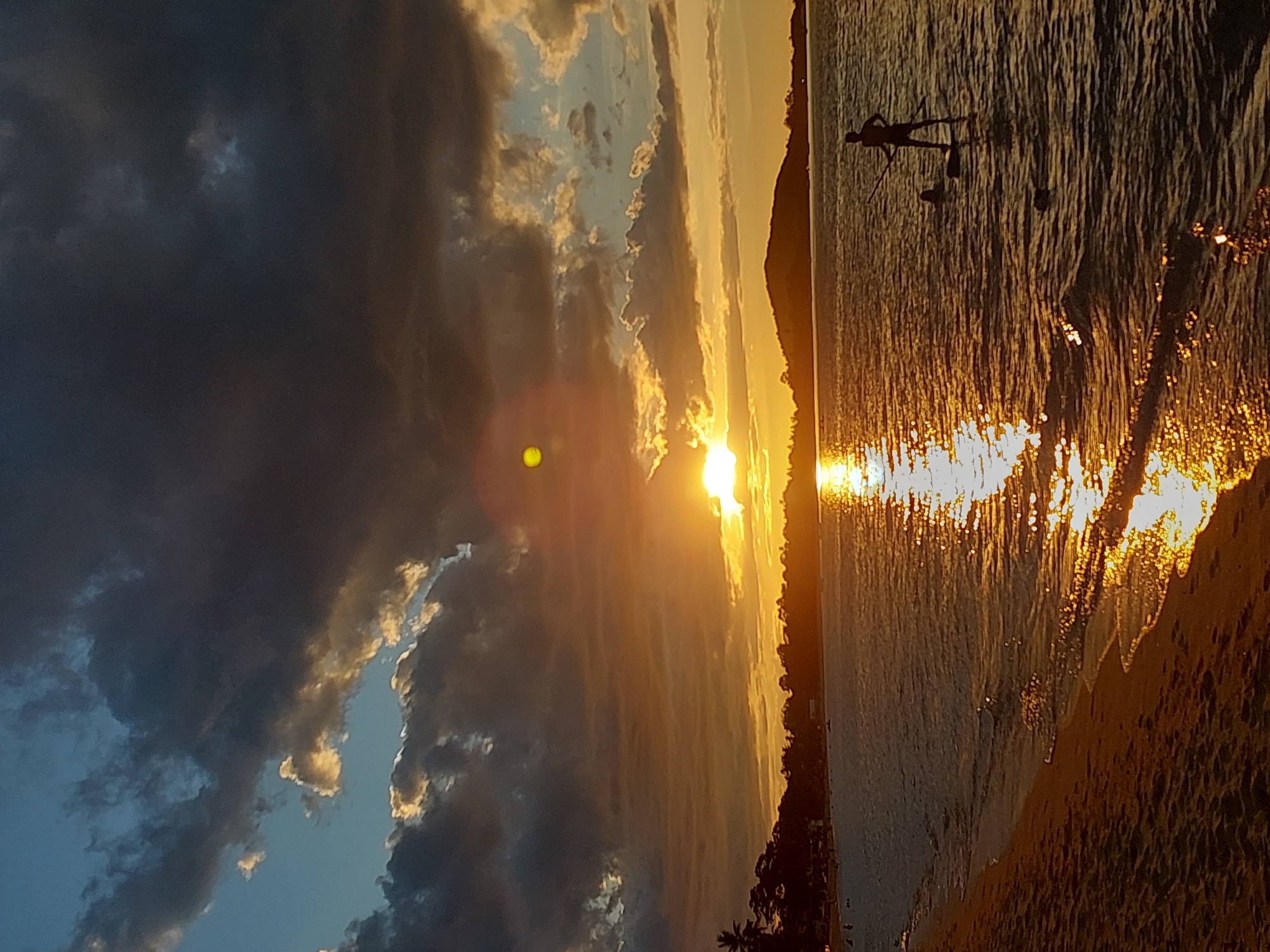 The image depicts a sunset over a beach in Baan Mae Nam, Thailand. The sun is low on the horizon, casting a warm golden glow across the scene. Dark, dramatic clouds dominate the left side of the frame, contrasting with the bright, sunlit sky on the right. The ocean is calm, reflecting the vibrant colors of the sunset, and small waves gently lap at the sandy shore.
In the water, a silhouetted figure is visible, appearing to stand on a paddleboard and holding a paddle. Another smaller silhouette nearby may be another person or an object. The beach itself is sandy, with some vegetation, including palm trees, visible along the shoreline, suggesting a tropical location. The overall mood is serene and tranquil, characteristic of a beautiful sunset. The time of day is clearly sunset, indicated by the low sun and the colors in the sky. The weather appears calm, with the sea being placid.