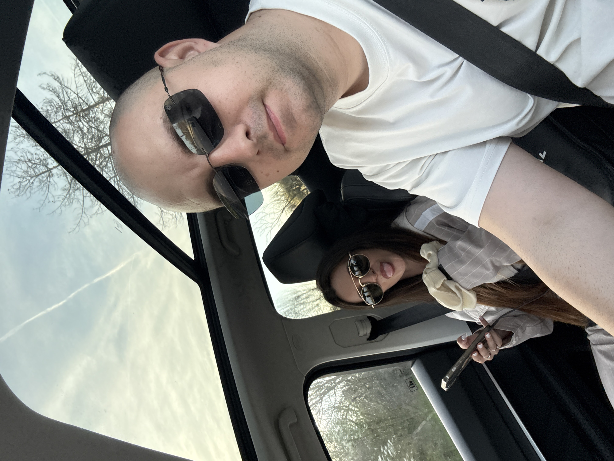 A selfie shows a bald man wearing a white t-shirt and sunglasses seated in a car. He is wearing a seatbelt. Behind him and below is a woman in sunglasses, sticking out her tongue, and holding a phone. A sunroof and car interior are visible. The location cannot be ascertained from this image.