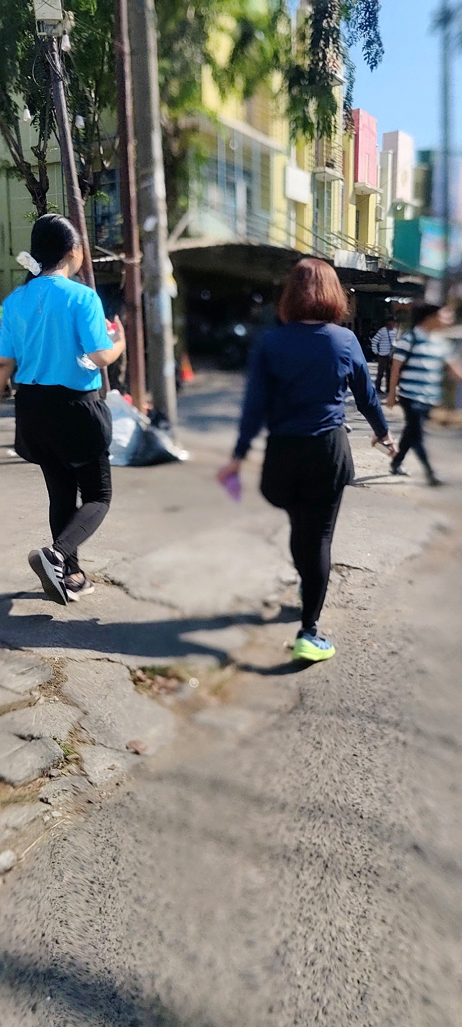 Two women are walking on a paved sidewalk in a city. The woman on the left is wearing a blue t-shirt and black leggings. She has her hair pulled back in a bun and is holding a water bottle. The woman on the right is wearing a dark blue long-sleeved shirt and black leggings. She has short, reddish-brown hair and is holding a purple object. In the background, there are buildings with colorful facades, trees, and utility poles. The ground is cracked and uneven in places, with some puddles of water. The scene appears to be in a commercial area. The image is blurry and out of focus, making it difficult to discern fine details. The lighting suggests it is daytime.
