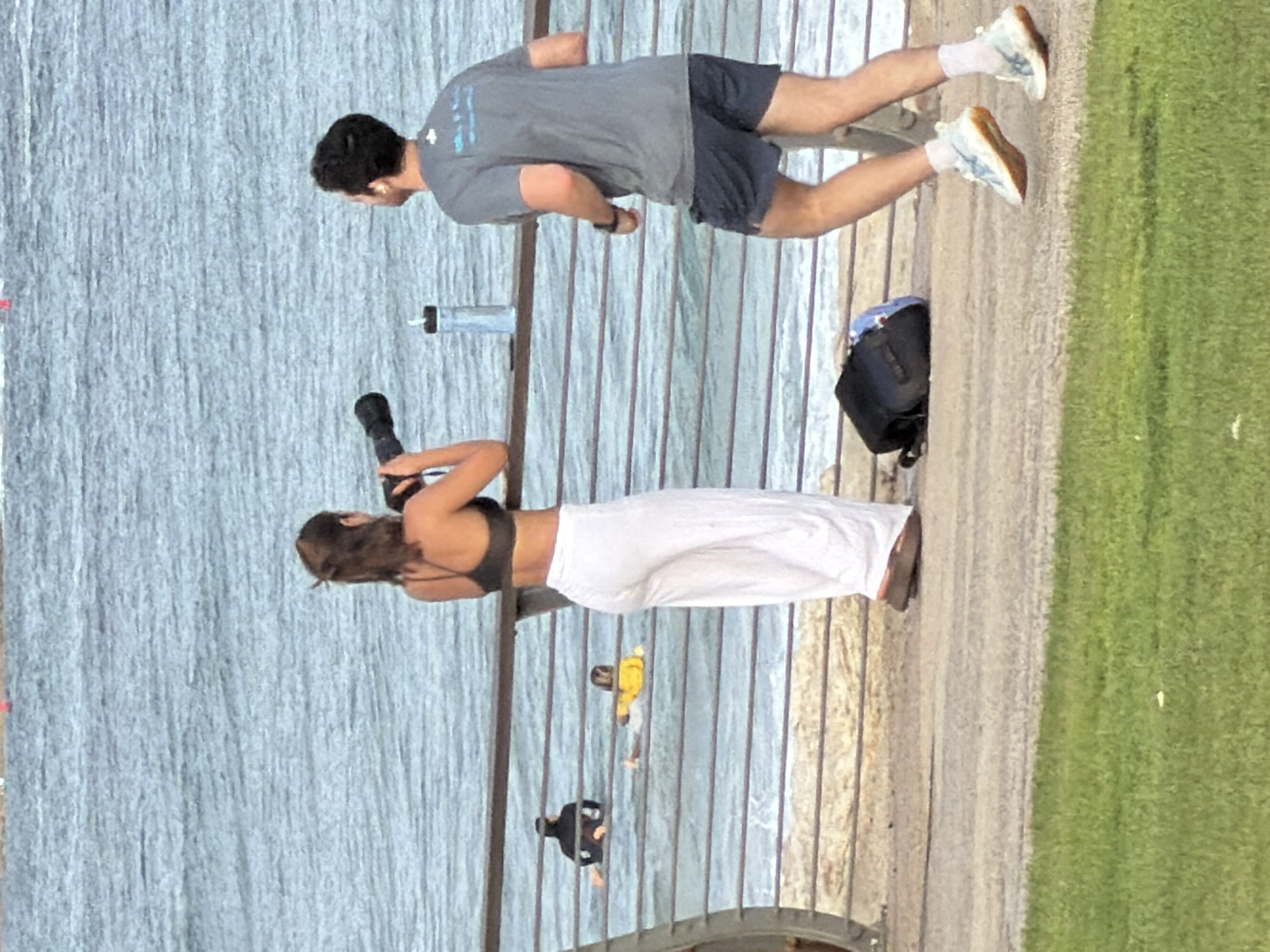 This media file captures an outdoor scene with several people. In the foreground, a woman is taking a photo with a camera. She is wearing a black bikini top and a long white skirt. Behind her, a man is walking, wearing a grey t-shirt and dark shorts. He is also wearing white sneakers. Further in the background, two other individuals are visible. One is wearing a yellow jacket, and the other is wearing a black top.
The setting appears to be a paved walkway next to a large body of water, likely the Mediterranean Sea, given the Tel Aviv context. Beyond the walkway and water, there is a grassy area. The lighting suggests it is daytime, possibly late afternoon due to the warm tones in the grass. The overall atmosphere seems relaxed, with people enjoying the outdoor environment. There are no visible text elements in the image.