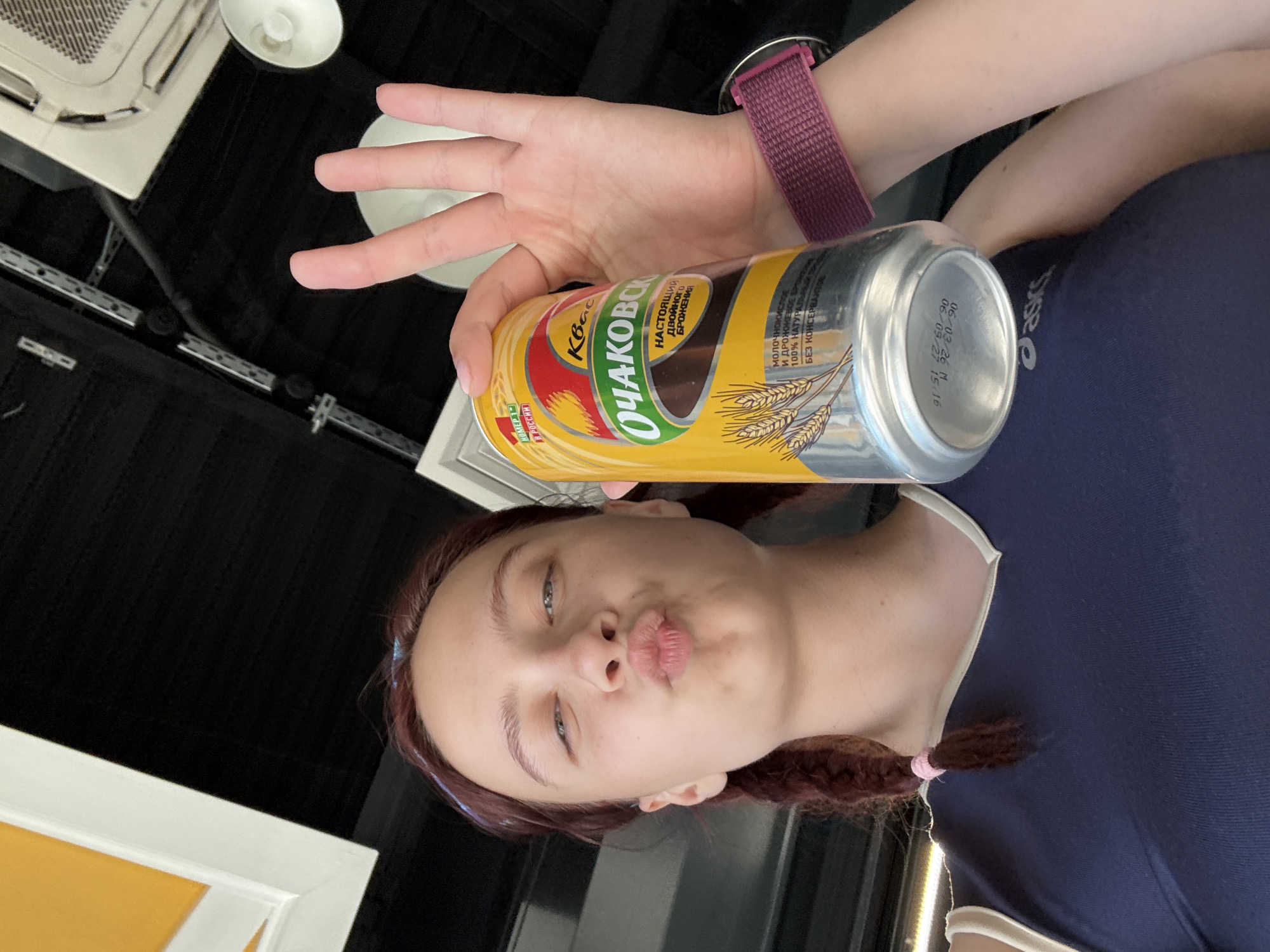 A girl is holding a can of "Ochakovo" kvass in her hand and puckering her lips. The can is yellow with images of wheat, and the text is in Russian. The girl is wearing a dark blue t-shirt with white trim and a pink hair tie on one of her braids. She is also wearing a maroon watch band. The environment includes a black ceiling, a white air conditioning unit, and a lamp. The OCR text on the can indicates that it is "Number 1 in Russia" and is a "100% natural product without preservatives". No location information about Казань, Russia can be extracted from this image.