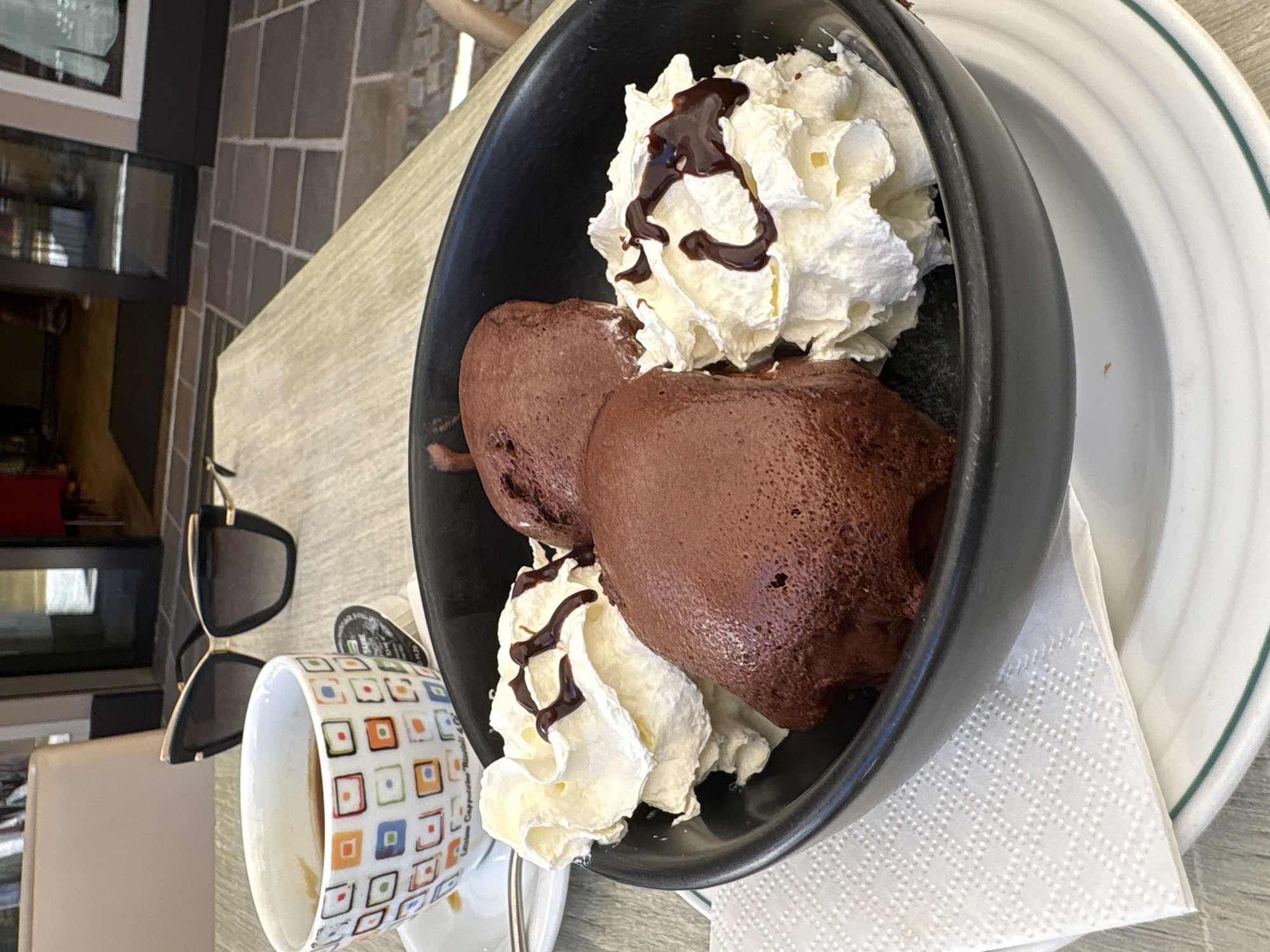 An overhead, eye-level shot shows a black bowl filled with chocolate mousse and whipped cream, resting on a white plate with a green rim. Three mounds of chocolate mousse are nestled among dollops of whipped cream, drizzled with chocolate sauce. To the left, a white ceramic cup with a colorful square pattern contains a dark liquid, likely coffee. A pair of black cat-eye sunglasses with gold accents rests behind the cup. The scene is set on a light-colored, textured table, with a blurred background suggesting an outdoor cafe setting in the city of Calfosch - Kolfuschg - Colfosco, Italy, indicated by the cobblestone flooring visible.