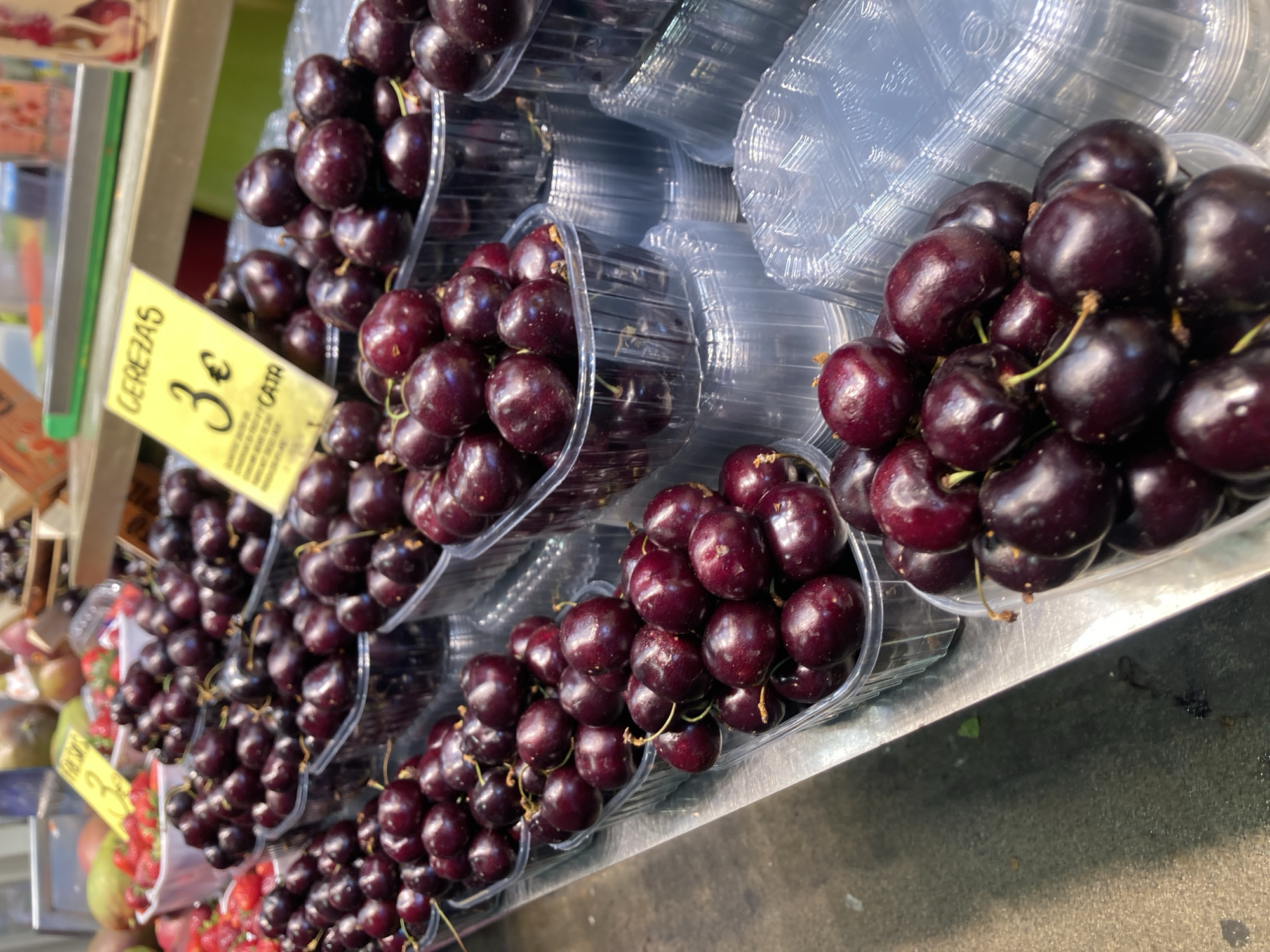 In Barcelona, Spain, a market stall displays an abundance of fresh cherries. The cherries are a deep, rich red, packed tightly in clear plastic containers, stacked in rows. A yellow sign with black text reads "CEREZAS 3€ CAJA," indicating the price and packaging type. The market setting is evident with other fruits visible in the background, such as strawberries and possibly mangoes, suggesting a vibrant food market atmosphere. The overall scene is focused on the fresh produce, highlighting the cherries as the primary subject.