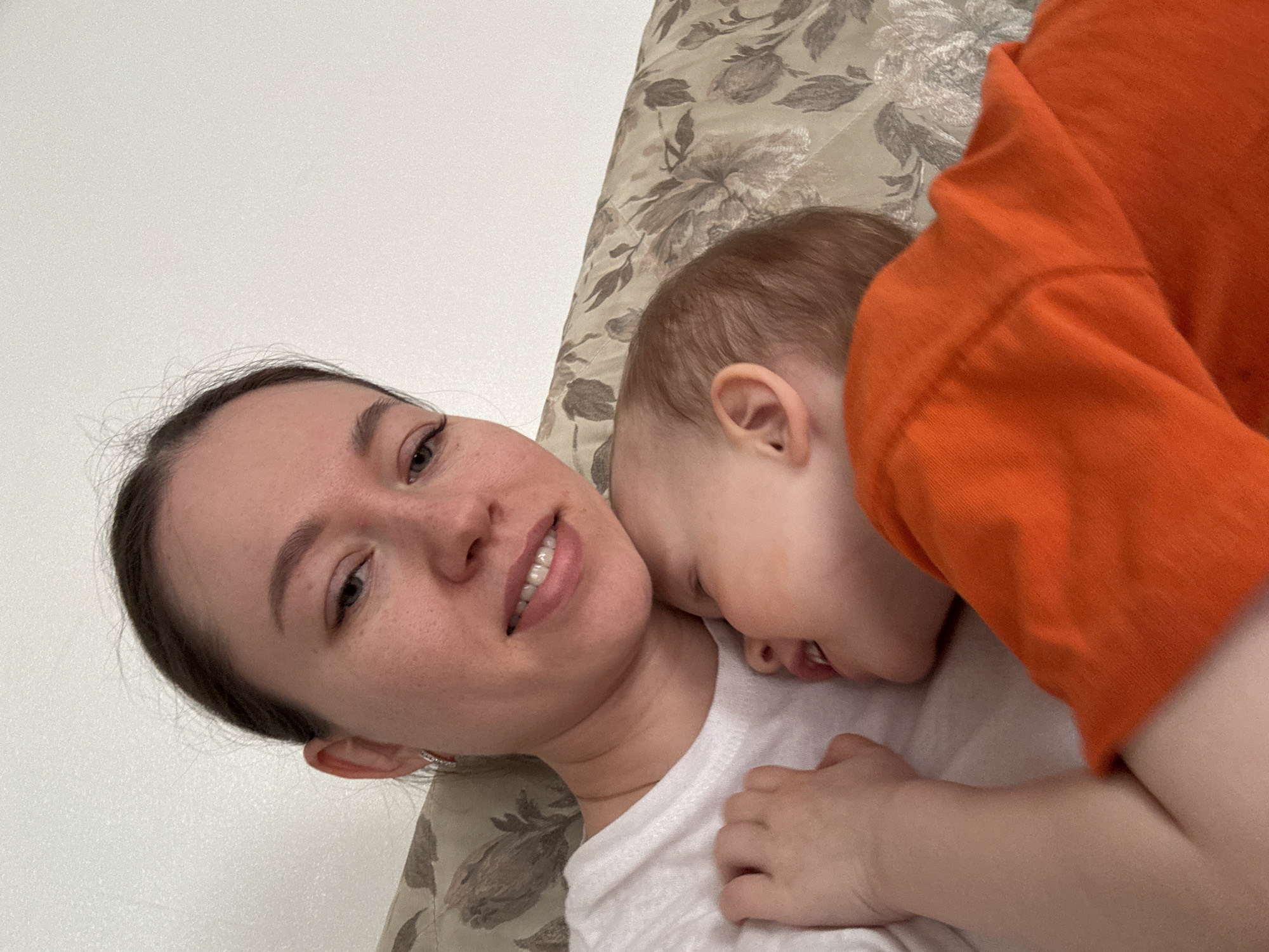 A woman is lying on a bed with a child. The woman is fair-skinned and has brown hair pulled back from her face. She is wearing a white shirt. The child, who is wearing an orange shirt, is lying on the woman's chest. The bed is covered in a floral patterned fabric. The location is Ферма, Russia.
