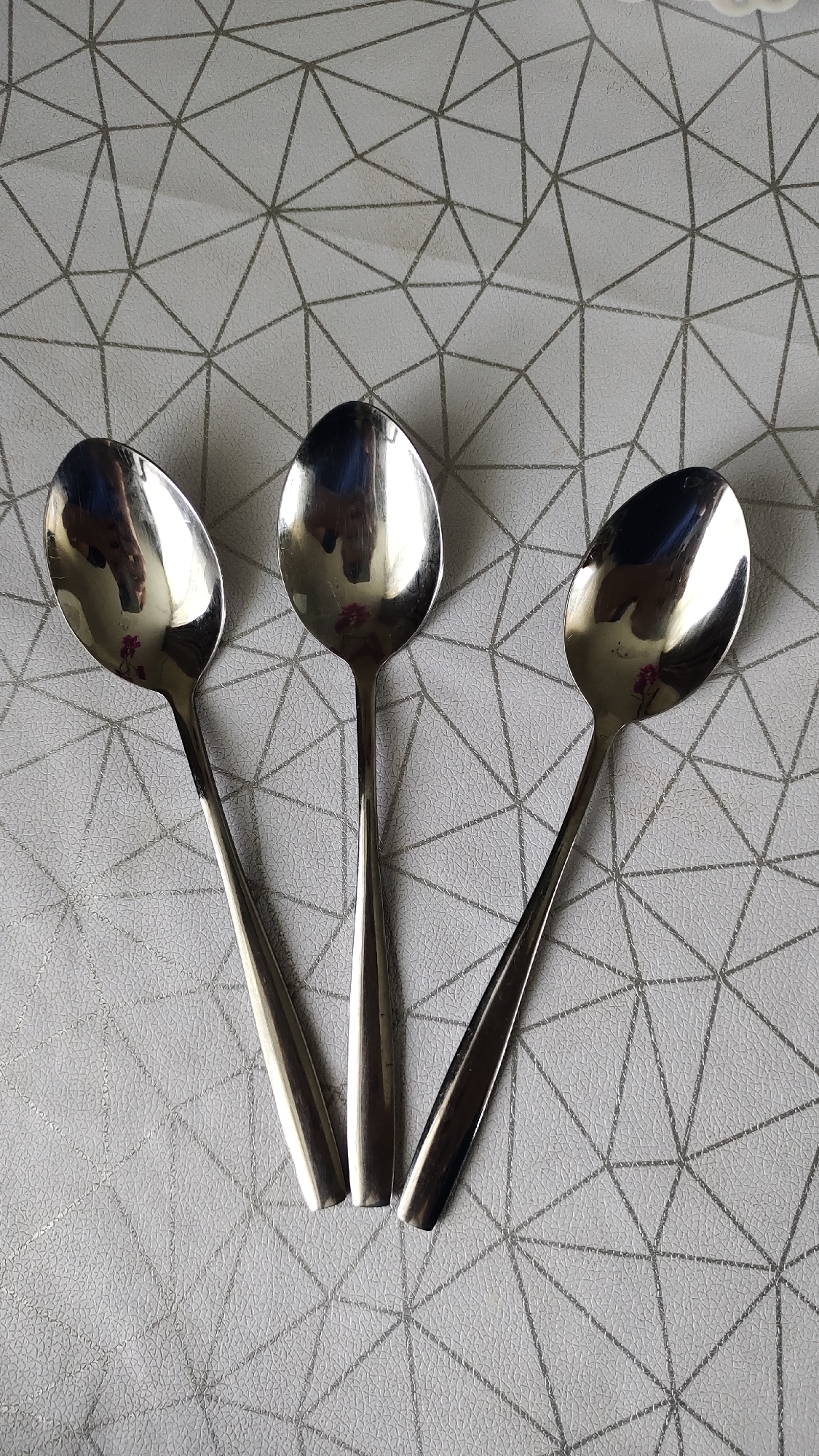 Three polished metal spoons are arranged in a fan shape on a light gray surface with a geometric pattern of intersecting lines. The spoons are positioned with their handles pointing downwards and their bowls upwards and to the left. The surface reflects the overhead lighting and the spoons, showing distorted images of what appears to be the ceiling and possibly the photographer. The spoons are clean and appear to be unused. The location is Unknown, Unknown.