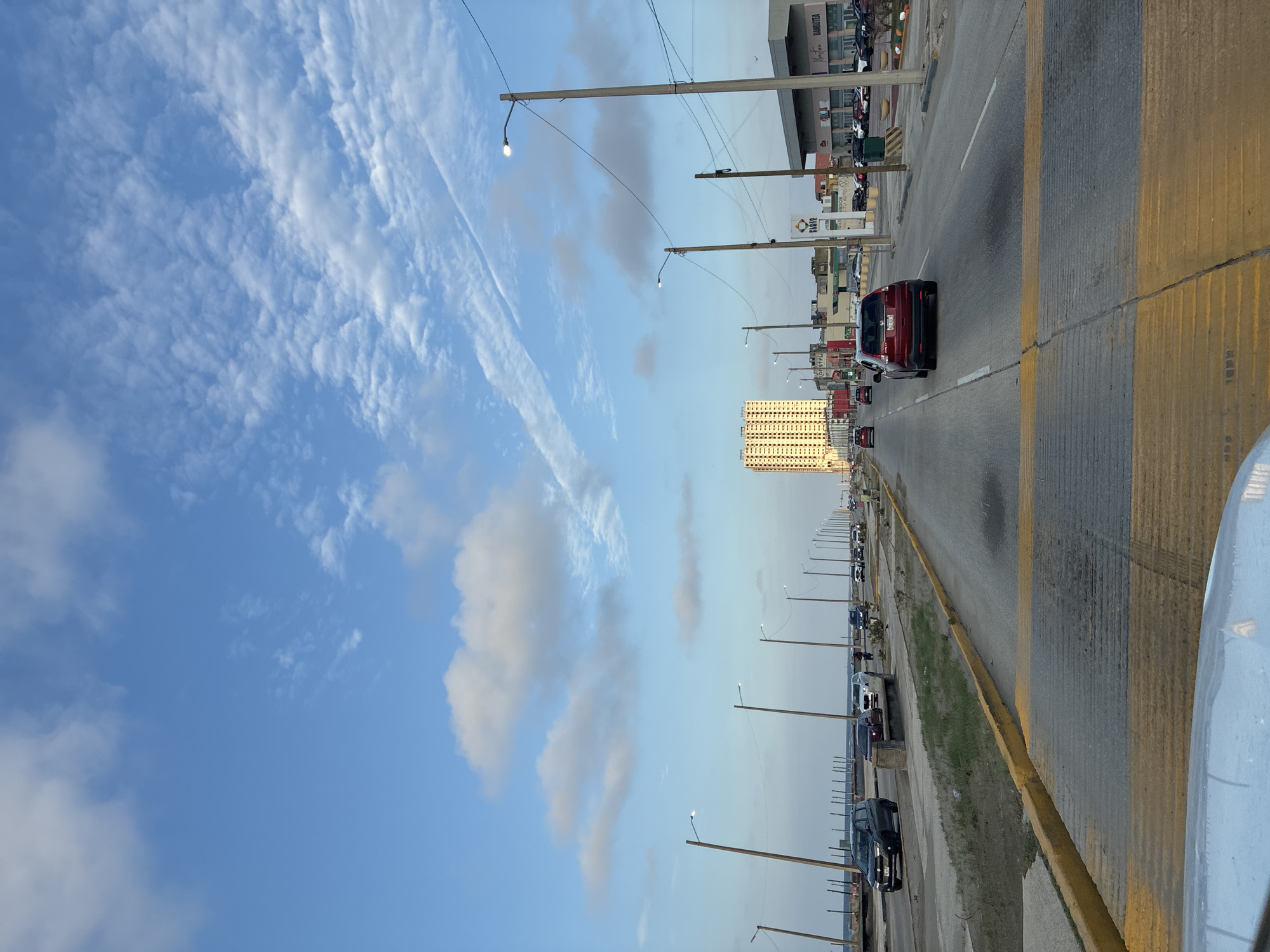 The image shows a road scene in Coatzacoalcos, Mexico. The view is from a car moving along a highway. The road has multiple lanes with yellow dividers. Several cars are driving in the same direction. On the side of the road are street lights and parked cars. Buildings, including a high-rise apartment or office building, are visible along the highway. The sky is blue with scattered clouds.