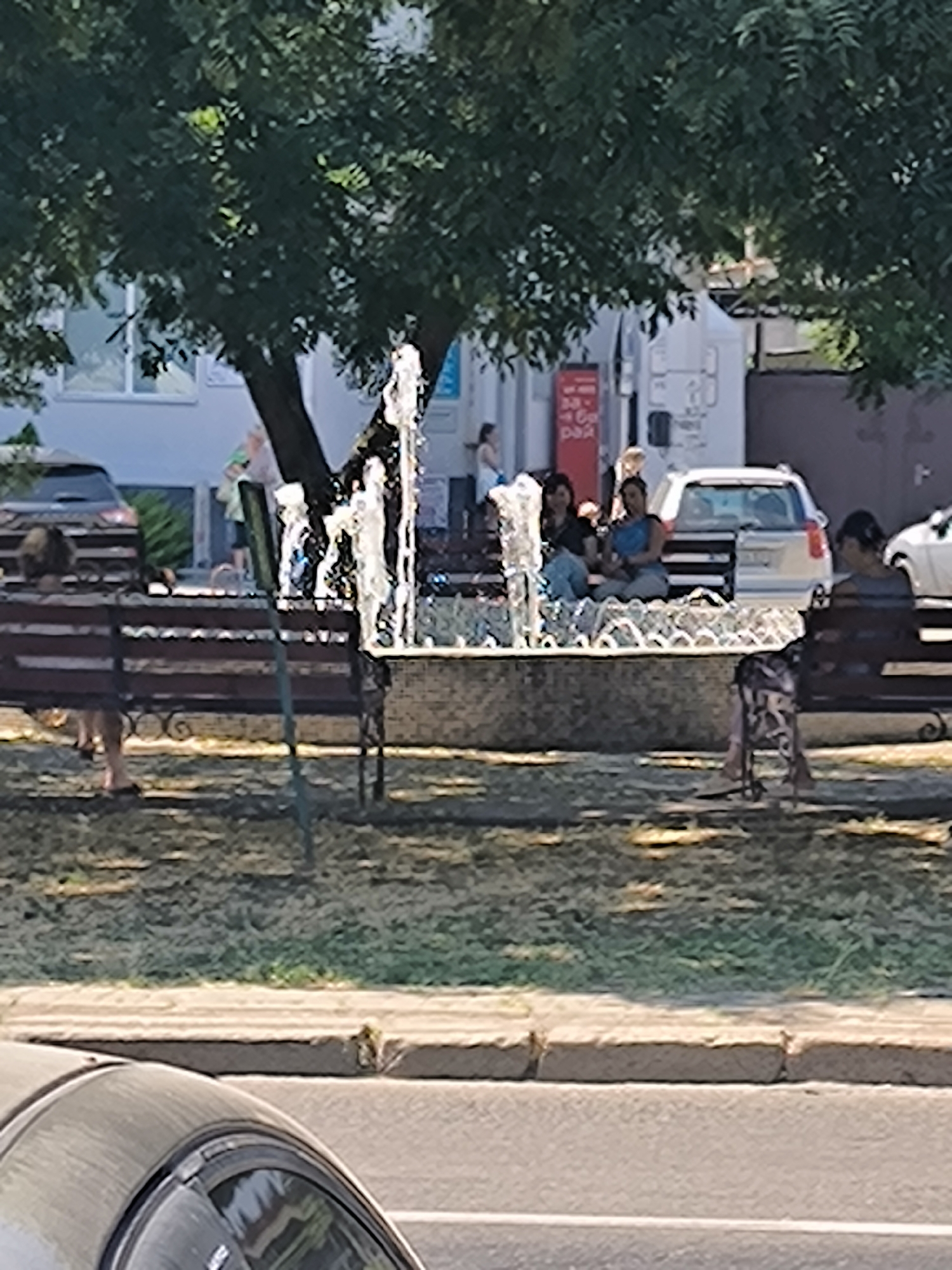 The image captures a scene in Mykolaiv, Ukraine, on a sunny day, likely in a park or public square, with a focus on a fountain and people relaxing on benches. In the foreground, the upper curve of a dark-colored car is visible, suggesting it is parked on a street adjacent to the area.
The central element is a tiered fountain with water spraying upwards, creating a dynamic visual. Surrounding the fountain, there are several park benches. On one bench to the left, a person with dark, short hair is seated, facing away from the camera, with their legs visible. Another bench, slightly further back and to the right of the fountain, accommodates three individuals. Two women are seated close together on the left side of this bench; one has dark hair and is wearing a dark top and light-colored pants, while the other has darker hair and is wearing a blue top and light pants. A third person, a male, is seated to their right, wearing a blue top and light pants. In the background, a white car is parked near a building.
To the far right of the fountain, another bench is occupied by a person seen from behind, wearing a light-colored top and dark bottoms. Their feet and legs are visible, and a backpack or bag rests on the ground beside them. Behind this bench, more people are visible in the distance, some standing and others sitting, under the shade of a large tree with dense foliage. The environment appears to be a well-maintained urban space, with grass and trees contributing to a pleasant atmosphere. The overall impression is one of a relaxed public setting, with people enjoying a break in the shade near the fountain.
