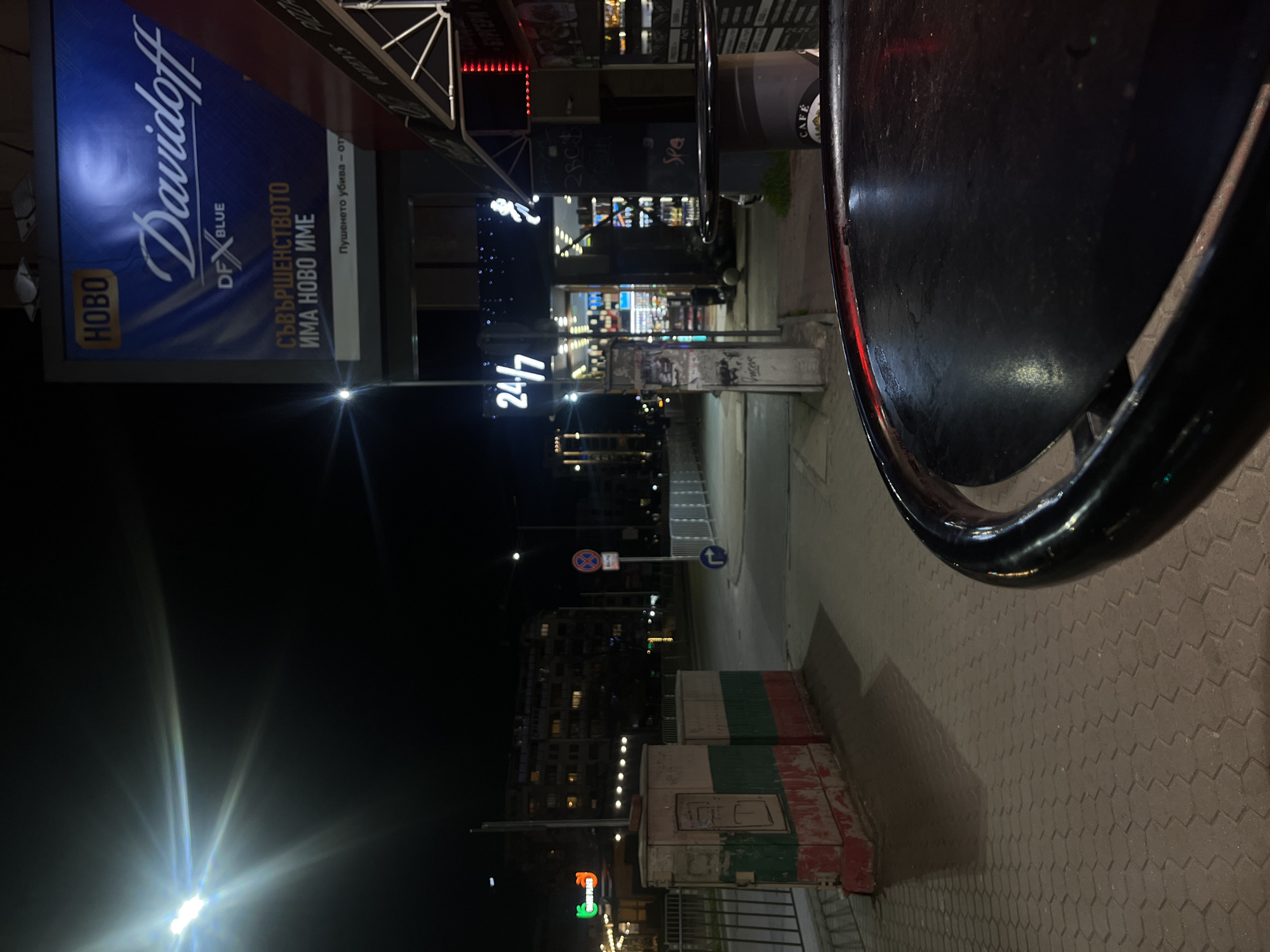 The image captures a night scene in Sofia, Bulgaria, showcasing urban elements. A partially visible black table dominates the foreground, with a nearby ashtray. In the midground, a sidewalk paved with zigzagging tiles and a low barrier painted in the colors of the Bulgarian flag are visible. Further along, there are illuminated signs, including one for "24/7," and advertisements, such as one for "Davidoff" cigarettes. Behind the signs, a street scene unfolds with buildings lit up in the distance. The scene is urban and suggestive of a café or bar setting at night.