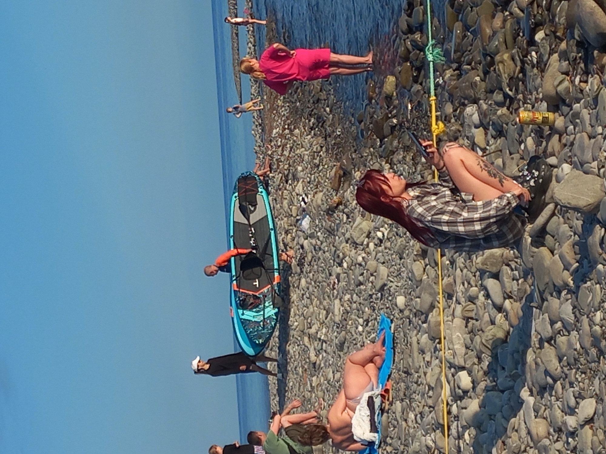 The image depicts a rocky beach scene, likely in Sochi, Russia, given the context. The sky is a clear, vibrant blue. In the foreground, a woman with long red hair is crouched down, looking at her phone. She is wearing a plaid shirt and shorts, and has tattoos on her legs. Beside her is a can of beer.
Further back, a woman in a pink top and shorts stands by the water. To her left, a young boy stands on a rocky embankment. Further left, two people are carrying a paddleboard. A woman in a black dress and white cap stands near them. To the left of the paddleboard, a person in a green shirt and shorts is seated on the ground. In the middle ground, a person is lying on a blue mat on the shore. Several other individuals are visible in the distance along the coastline. The sea stretches out towards the horizon. The beach is covered in grey, rounded stones of various sizes.