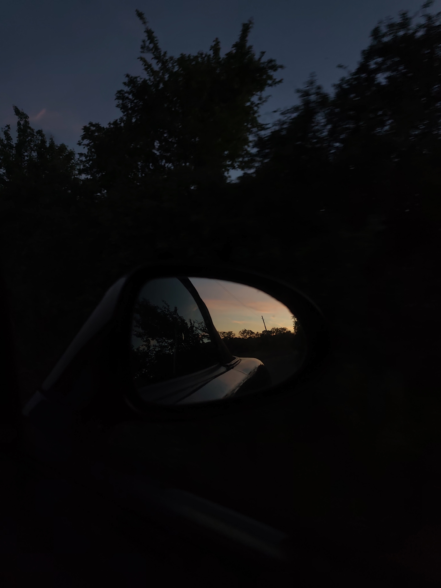 A side mirror of a vehicle reflects a sunset with a glimpse of trees and a utility pole in the distance. The surrounding environment outside the mirror is dark with blurry trees visible against a twilight sky. The scene appears to be taken from inside a vehicle at dusk. Location: Unknown, Unknown.