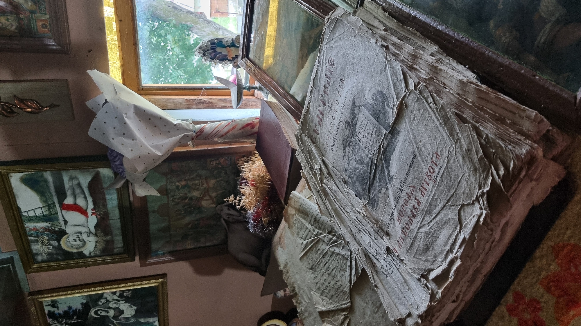 This media file captures a cluttered, indoor scene, likely a room in Vyshka, Ukraine. The focus is on a large, aged stack of what appears to be newspapers or old documents, piled precariously on top of a book. The paper is yellowed and worn, with visible tears and creases, suggesting significant age and frequent handling. Some of the documents have text and images printed on them, though the details are mostly obscured by the stack's condition.
To the left of the stack, a window with wooden frames looks out onto a greenery-filled outdoor environment, suggesting daytime. Sunlight filters through the window, illuminating the room and casting shadows. Several framed artworks hang on the wall. One painting, towards the left, depicts a figure that resembles a child, possibly a religious depiction, with a red sash. Another painting further to the right shows a more complex scene with multiple figures. A bouquet of dried flowers and other decorative elements is tucked beside the stack of papers.
A white wrapping with polka dots covers a portion of something on the windowsill, potentially a gift or another item. The overall impression is one of accumulated possessions, perhaps in a storage area or a room filled with historical items. The lighting suggests it is daytime, and the view from the window hints at a natural, possibly rural, setting outside. No specific activity or emotion is clearly depicted, but the aged nature of the objects could evoke a sense of history or nostalgia. Some text is visible on the topmost newspaper, including what appears to be Cyrillic script, but it is too small and blurry to read clearly.