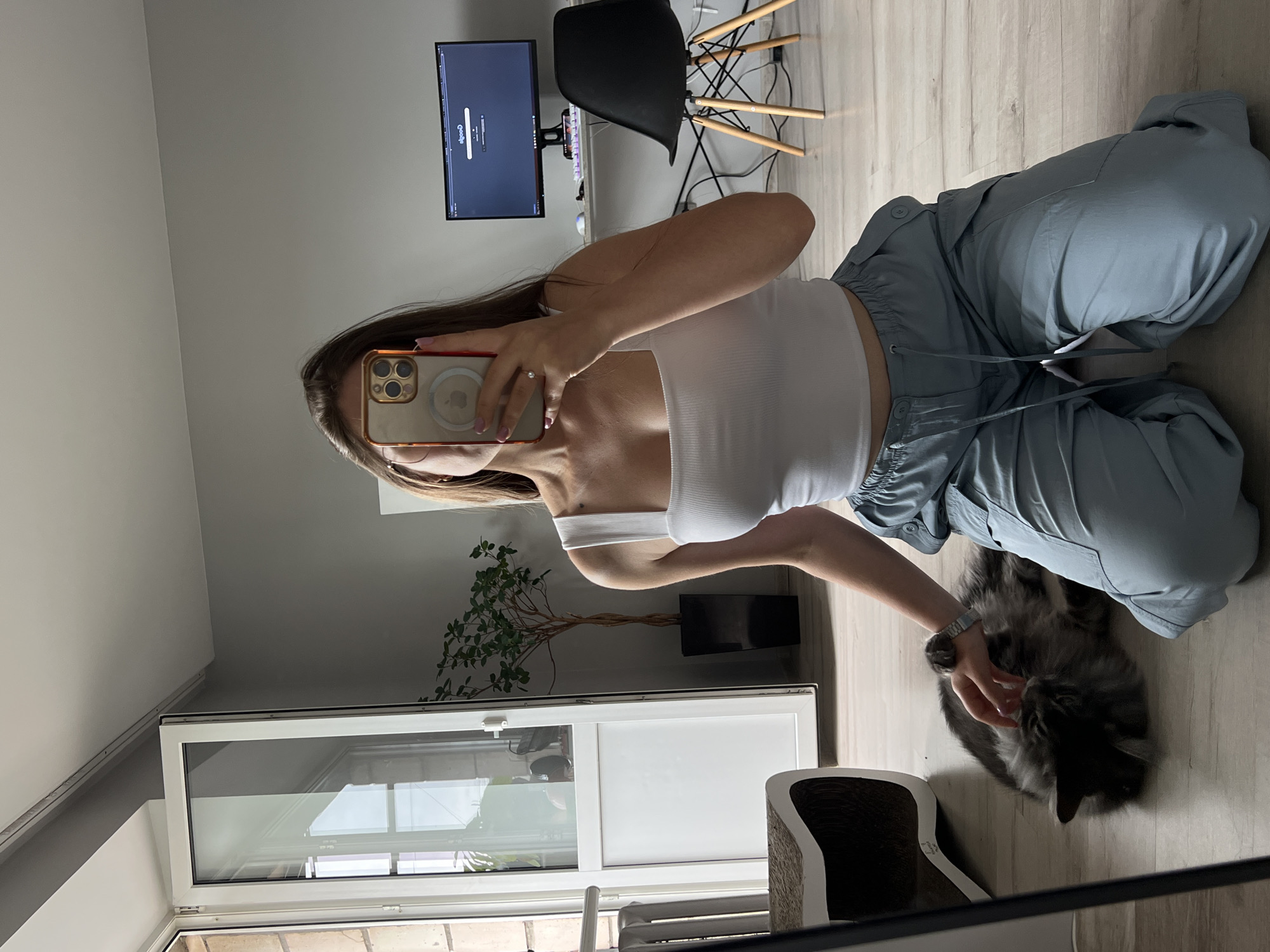 A woman is taking a mirror selfie while kneeling on the floor in a room. She is wearing a white tank top and light blue cargo pants. A gray cat is lying on the floor next to her, and she is petting it. To the left of the woman is a cat scratching post. Behind her, a desk with a computer monitor, keyboard, and chair is visible. A plant in a black pot is next to a white door on the left side of the room. The overall environment suggests an indoor living space, possibly an apartment in Ternopil, Ukraine, given the presence of a standard balcony door.