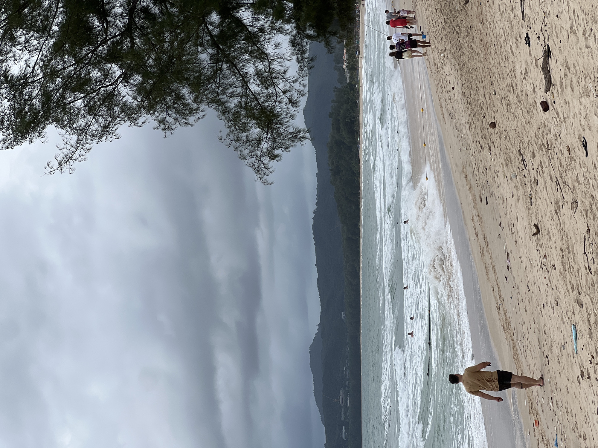 The image depicts a beach scene under a cloudy sky, with rough seas and dark mountains in the background. In the foreground, a man in a tan t-shirt and black shorts walks away from the camera along the sandy shore. Further down the beach, a group of people, including women in white tops and a man in a white shirt and shorts, walk together. Some individuals are swimming in the wavy ocean, and a buoy line is visible in the water. The sand is littered with debris, including plastic, wood, and coconuts. A tree with dark green foliage hangs over the right side of the frame, obscuring part of the sky. The overall atmosphere is somber due to the overcast weather.