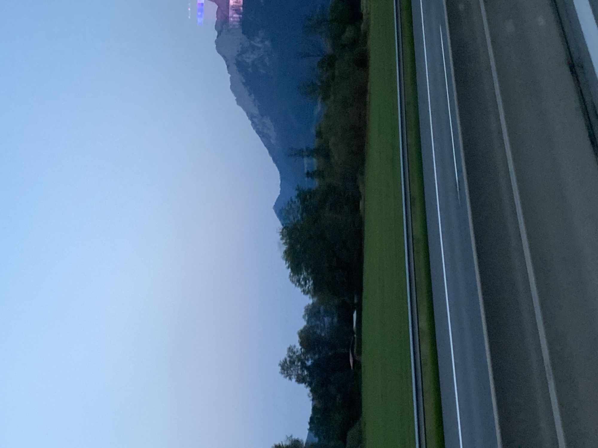 The image depicts a landscape scene with a highway in the foreground and mountains in the background. The sky is a pale blue, fading to a lighter shade near the horizon. The mountains are dark blue and jagged, with some lighter patches suggesting snow or rock. Trees of various shades of green and dark brown are scattered across the mid-ground, with a patch of bright green grass in front of them. A red-roofed structure is partially visible among the trees. The highway is asphalt, with white lines marking the lanes, and a metal guardrail separating it from the grassy area. The overall lighting suggests either early morning or late evening. There are no people or animals visible in the image. The location is identified as the city of Oberaudorf, Germany.