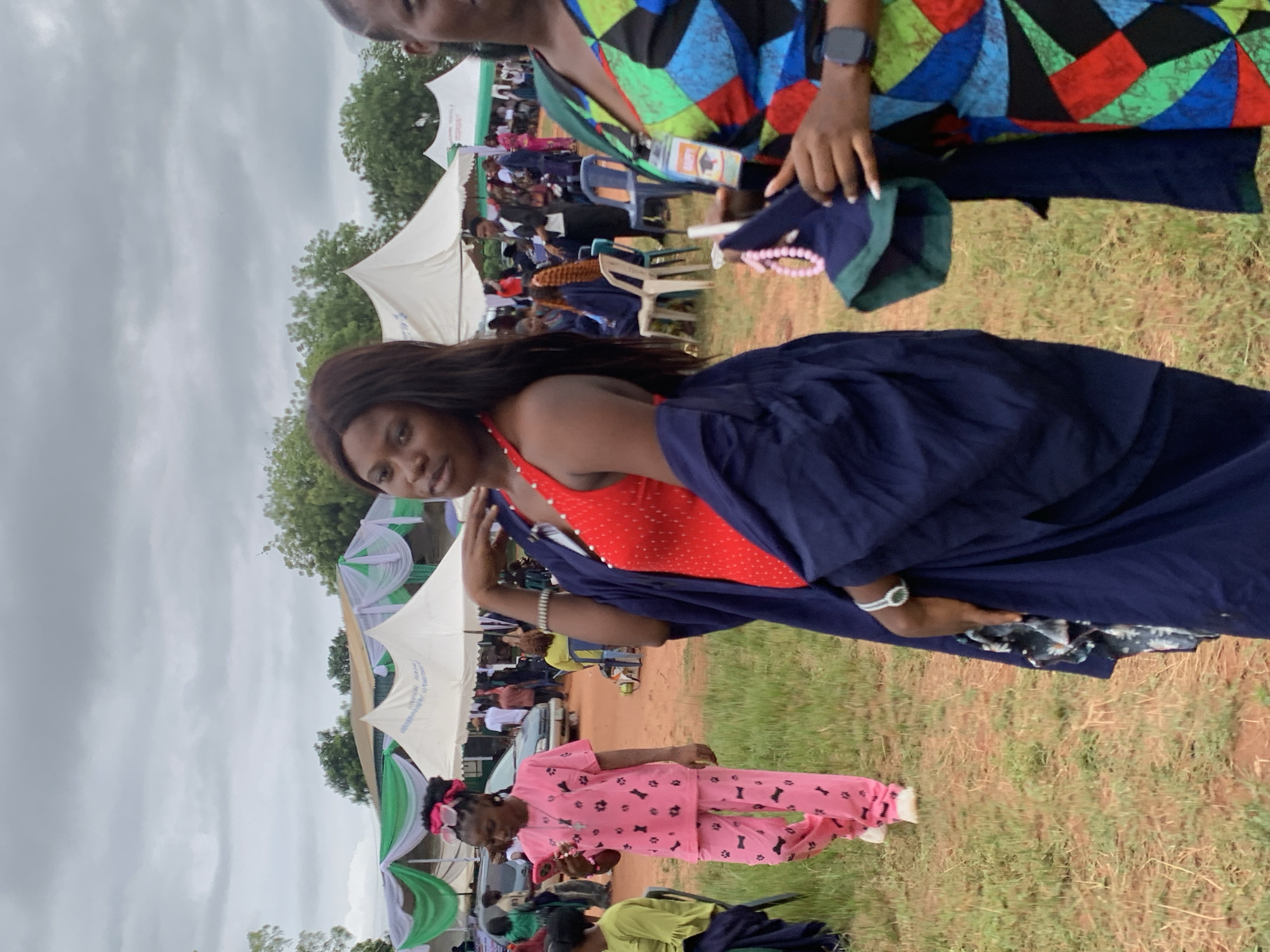The image shows an outdoor event in Ukwu-Abwa, Nigeria. A woman in a red dress and a dark blue gown stands in the foreground. Another woman with a colorful geometric-patterned top stands partially in the upper right corner, holding a beaded necklace and an identification card. In the background, there are tents decorated with green and white fabric, under which people are seated. A child in pink pajamas with paw print patterns walks in the foreground as well. The ground is covered in dry grass, and trees are visible in the background. The sky is overcast.