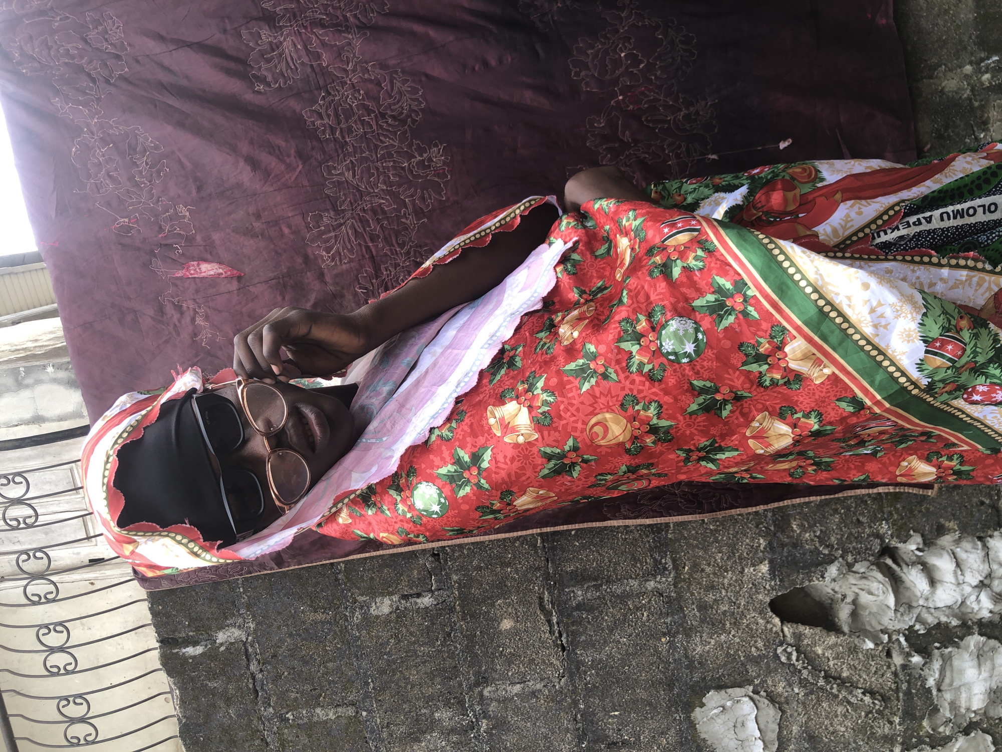 In Ado, Nigeria, a person is standing in front of a textured wall and a patterned cloth. The person is wearing a black headscarf, sunglasses, and a vibrant red garment adorned with Christmas-themed prints, including holly, bells, and ornaments. The person's right hand is raised, touching the sunglasses. The background features a weathered concrete wall on the left and a dark, patterned fabric on the right. A metal fence is visible above the wall. The overall lighting suggests an outdoor setting.