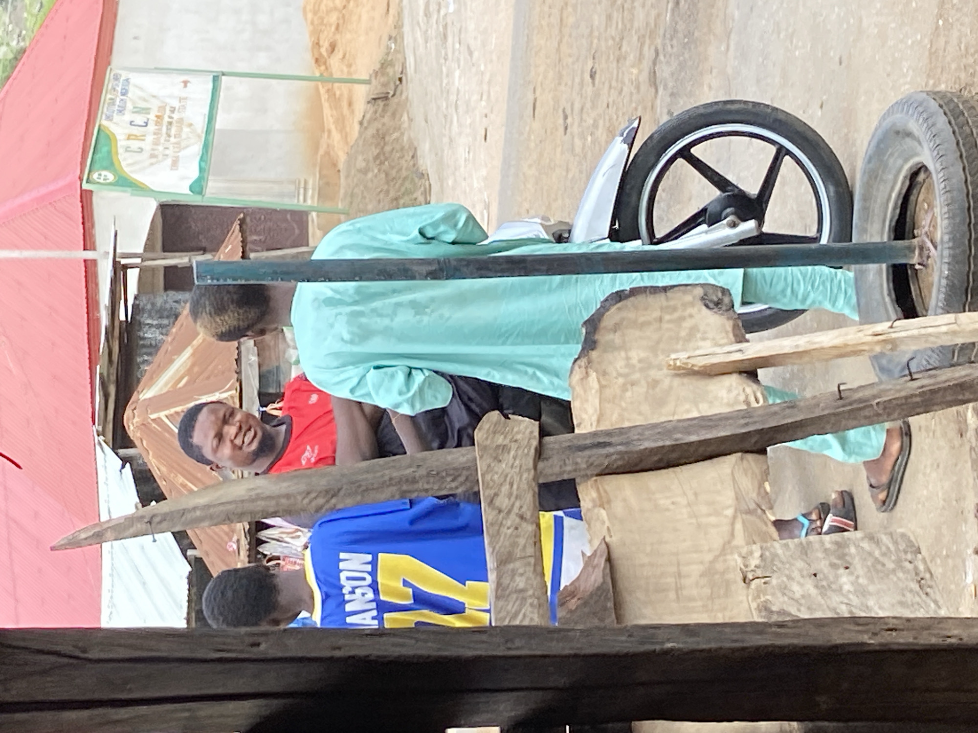 In Mararaba, Nigeria, a medium shot depicts a scene featuring several individuals and objects. Two men are visible; one wears a red shirt, is smiling, and appears to be looking towards the viewer. Another man is wearing a blue shirt with the name "ANSON" and the number 27 on the back. A person in a light blue garment stands adjacent to what seems to be a makeshift trolley with wheels, constructed using wood and metal components. The trolley has two wheels, one of which is a car tire. The scene is partially obstructed by wooden structures in the foreground, suggesting the perspective of an observer viewing from within or behind a shelter. In the background, there's a building with a red roof and a sign, though details on the sign are indistinct. The surroundings suggest a modest residential or commercial area.