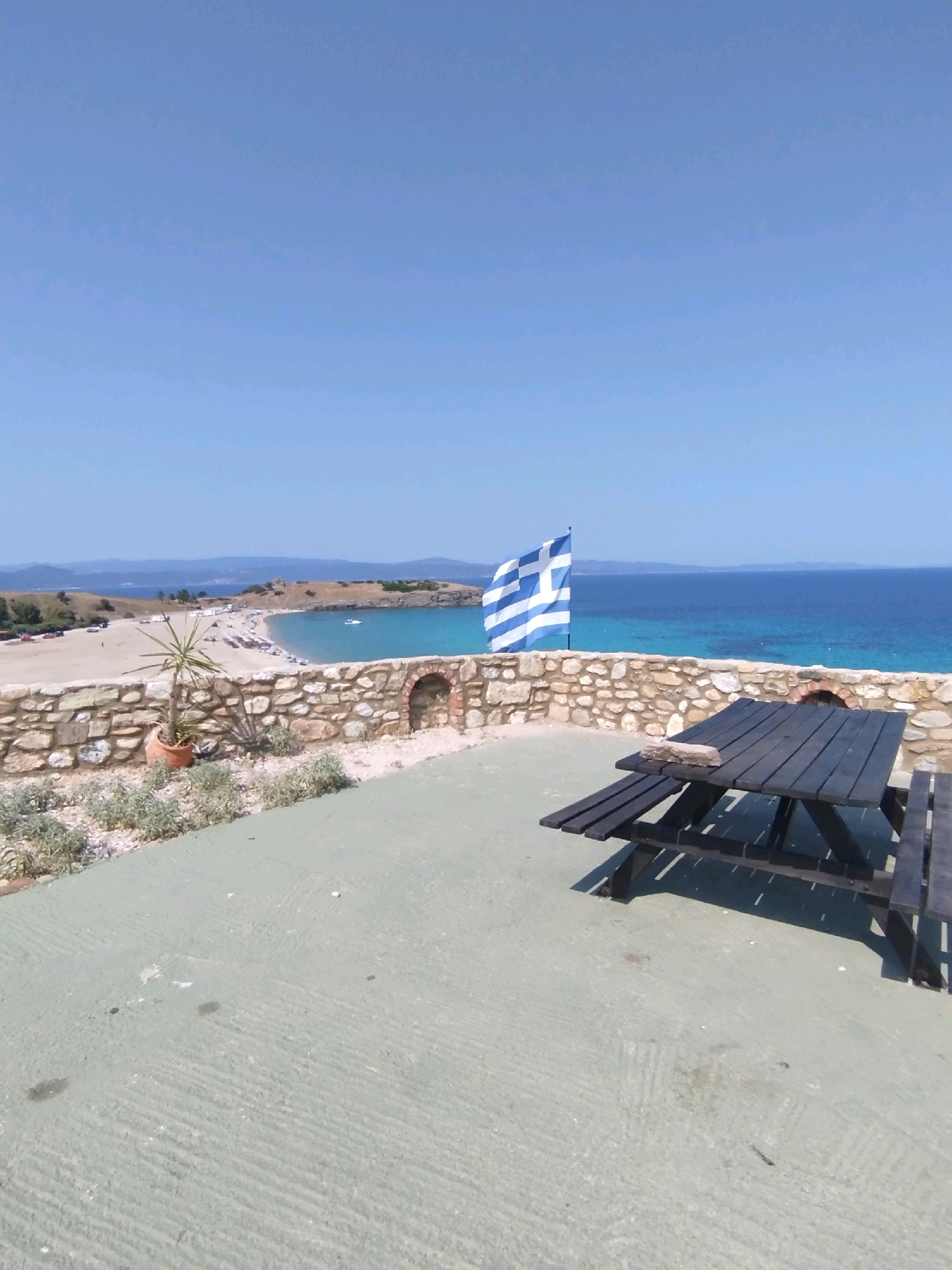 In Sithonia Municipal Unit, Greece, a wide, low-angle shot captures a sun-drenched outdoor seating area overlooking a coastal landscape. The foreground is dominated by a light green, textured concrete surface, with a dark wooden picnic table and bench set in the middle right of the frame, casting sharp shadows. A grey stone wall, constructed with irregular shapes, runs across the midground, forming a barrier and featuring an arched opening. To the left of the wall, a potted plant with spiky leaves stands near a patch of low-lying greenery.
Beyond the wall, a sandy beach is visible, dotted with numerous white beach umbrellas and some scattered people. The sea is a vibrant turquoise blue, transitioning to a deeper blue further out, with distant hills and islands forming the horizon under a clear, pale blue sky. A white boat is anchored in the water near the beach. A Greek flag, blue and white, flutters prominently on a flagpole behind the stone wall, adding a national emblem to the scene. The overall impression is of a peaceful, sunny day at a seaside location.