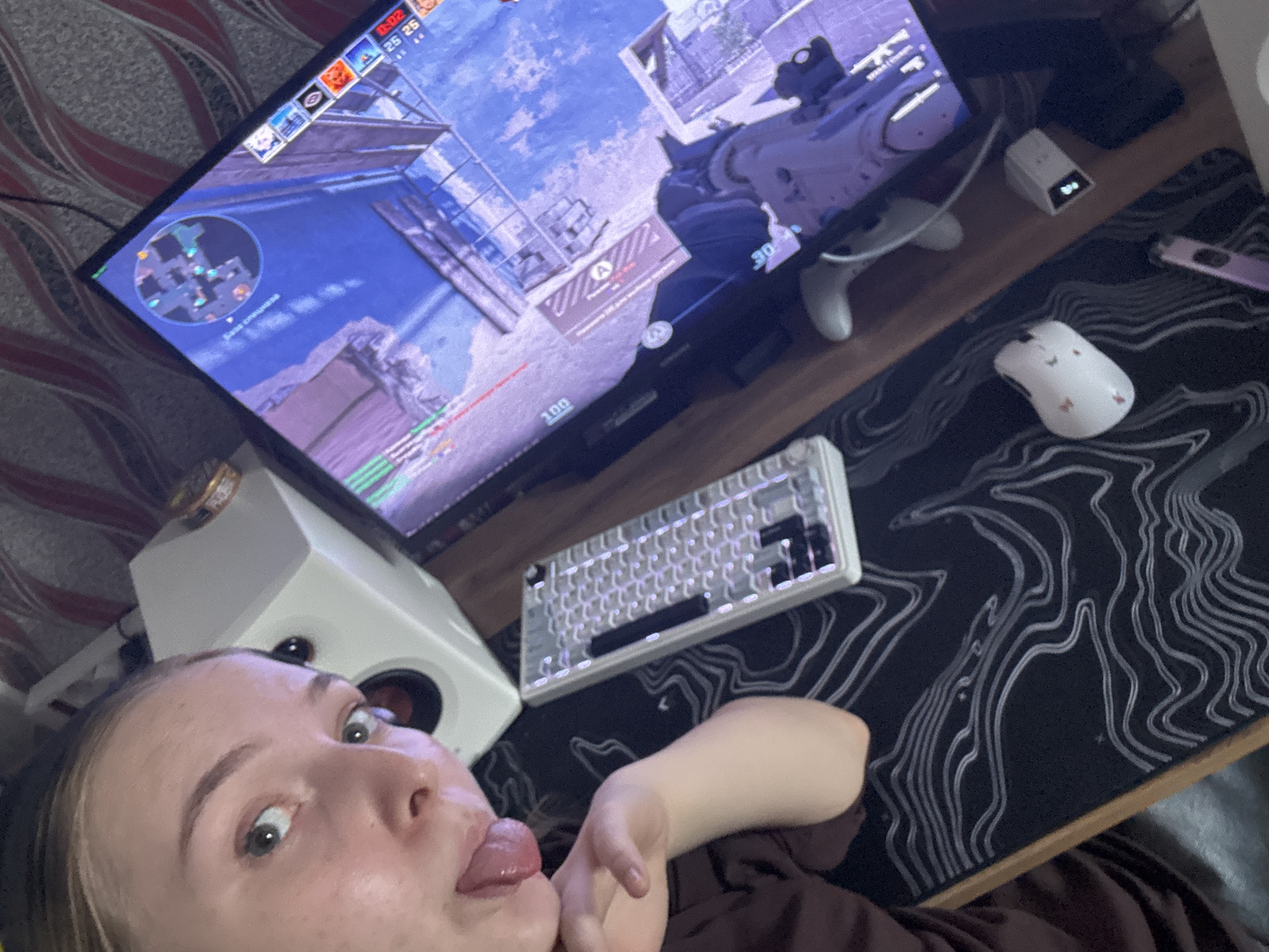 A close-up, high-angle shot depicts a woman's face, with her tongue sticking out, positioned near a gaming setup. A computer monitor displays a first-person shooter game, complete with a minimap in the corner, health/ammo indicators, and in-game action. Below the monitor is a gaming keyboard with white keycaps. To the right of the keyboard is a gaming mouse, also white with butterfly designs. A patterned mousepad lies beneath the keyboard and mouse. A white speaker is positioned near the woman's face, partially obscured. A game controller sits near the monitor. The location is indeterminate; there is no information to place it in Perm, Russia.