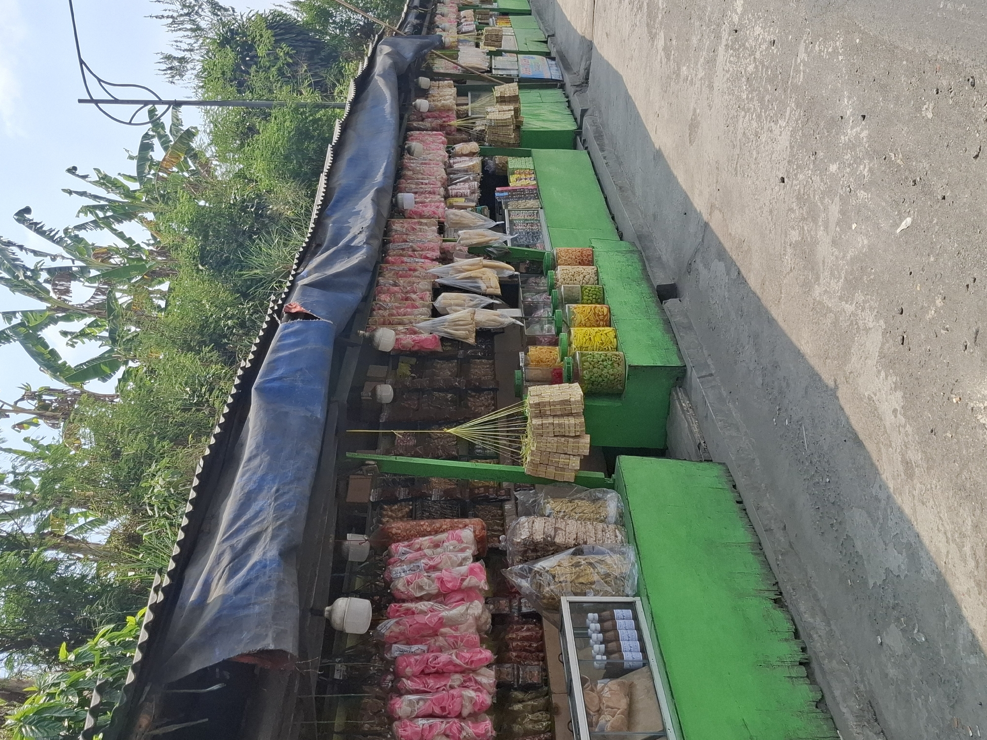 This is a photograph of an outdoor market stall in Pamuruyan, Indonesia. The stall is long and narrow, with a corrugated metal roof covered by a dark blue tarp. Green wooden structures form the base of the stall, displaying various packaged food items. Hanging from the roof are numerous bags of snacks, including pink and white crackers and other dried goods. In front of the stall, on the green wooden base, are several clear jars filled with colorful snacks, as well as a glass display case containing bottles of sauce. The ground in front of the stall is a paved pathway or road, and a shadow from the stall stretches across it. In the background, lush green vegetation and trees are visible, with a clear sky above. The overall impression is of a typical roadside market in a tropical region.