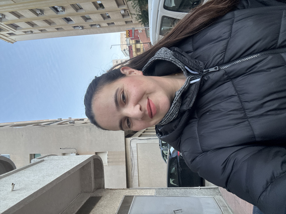 A woman takes a selfie outdoors in an urban setting. The image is rotated 90 degrees clockwise from its natural orientation.
The woman, appearing to be in her 20s or 30s, is the central focus. She has dark hair, mostly pulled back, with some strands visible around her shoulders. Her face shows a gentle, pleasant smile, with her lips closed and tinted with a soft pink or nude lipstick. She has light-colored skin with some faint freckles visible on her cheeks. Her dark eyes are directed slightly off-camera, contributing to a natural, candid expression. She is wearing a black puffer jacket with a slight sheen, and a gray, ribbed, possibly knit or fleece lining is visible at the collar and around the hood area, contrasting with the black main fabric. A metallic zipper pull is also visible on the jacket.
The background reveals a bright, clear blue sky with only a few faint wisps of clouds, suggesting a sunny day. Tall, light-colored (beige or off-white) multi-story buildings flank the sky area, featuring numerous windows or balconies and visible external air conditioning units.
At what would be ground level in a correctly oriented image, a street or alleyway is visible between buildings. A white van is partially seen behind the woman's right shoulder (when oriented correctly), and a black car is visible further down the street. Architectural elements such as curved concrete structures are present in the lower left foreground (when oriented correctly). A metal gate or door can be seen at ground level. In the distance, a yellow sign with black markings, possibly an arrow or symbol, is visible near a light pole, but no legible text can be discerned. The overall impression is one of a casual, daytime selfie taken in a city environment.