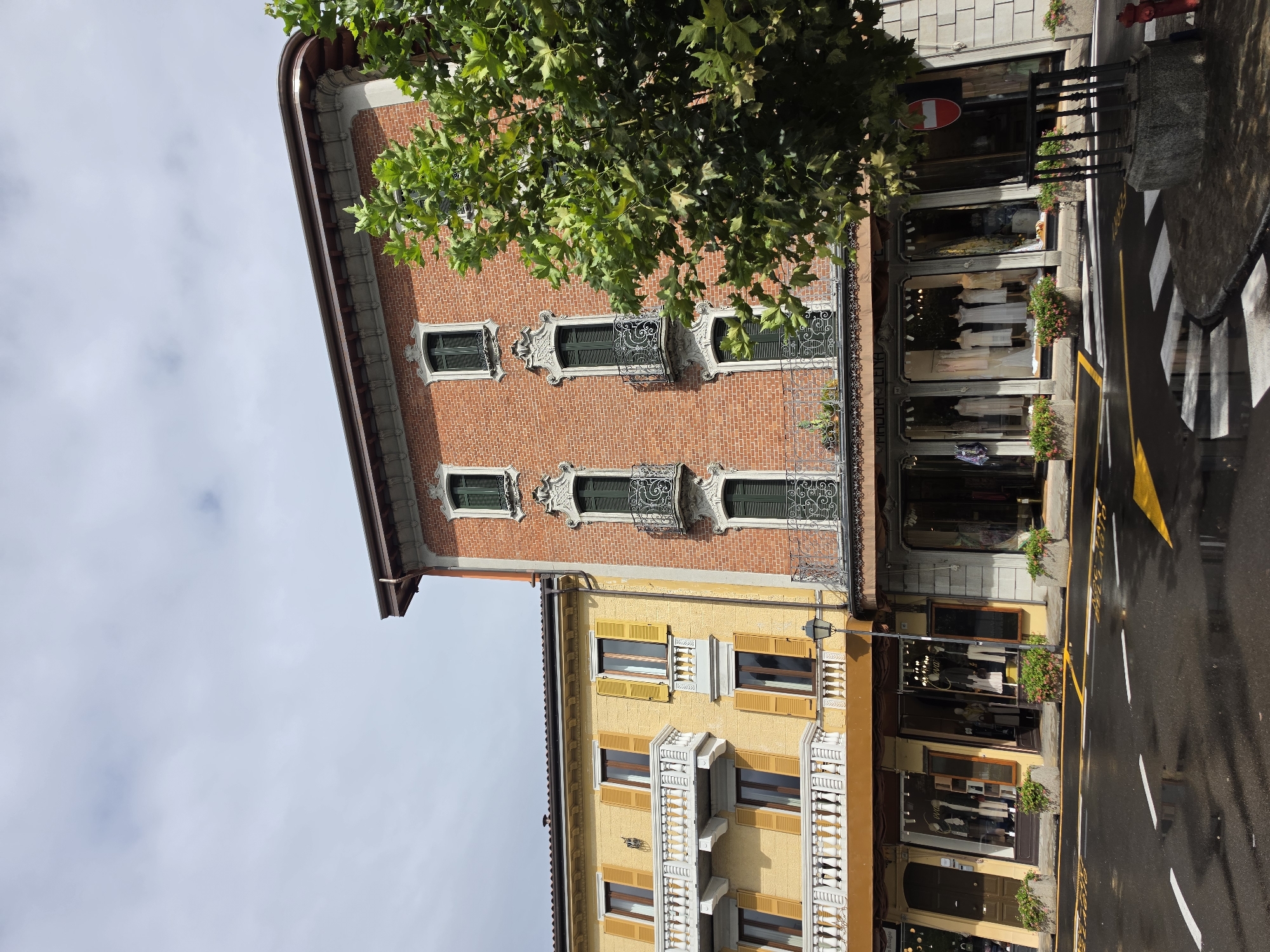 The image captures a street view in San Giovanni, Italy, featuring two adjacent buildings with shops on the ground floor and residences above. The building on the left is painted yellow and features balconies with white railings and yellow shutters on the upper windows. The building on the right is constructed of red brick with ornate white trim around the windows and decorative wrought-iron balconies. A lush green tree partially obscures the right side of the brick building.
The street in front of the buildings is wet, suggesting recent rain, with white and yellow road markings, including a pedestrian crossing. The shops display mannequins in formal attire through their large display windows. A street lamp stands between the two buildings. A red and white traffic sign is visible on the right side of the image. The sky above is overcast with scattered clouds. No people are visible in the frame. The overall scene depicts a quiet, European street on a potentially damp day.