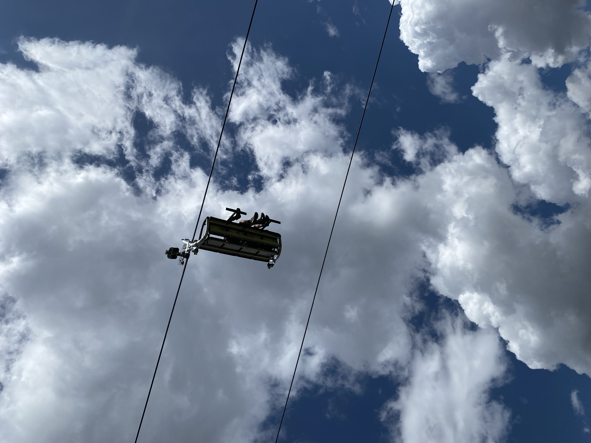 A single chairlift with two people is suspended in the air against a bright blue sky filled with fluffy white clouds. The chairlift is a metal structure designed to transport passengers and is attached to two overhead cables. The people in the chairlift are facing away from the camera and appear to be wearing casual clothing and shoes. One person is wearing shorts and the other is wearing pants. The chairlift is positioned slightly off-center in the frame, with the cables extending diagonally across the image. The overall impression is one of a clear, sunny day, possibly during a summer activity like hiking or visiting an amusement park. The image does not provide any specific clues about its location in the city of Unknown, Italy.