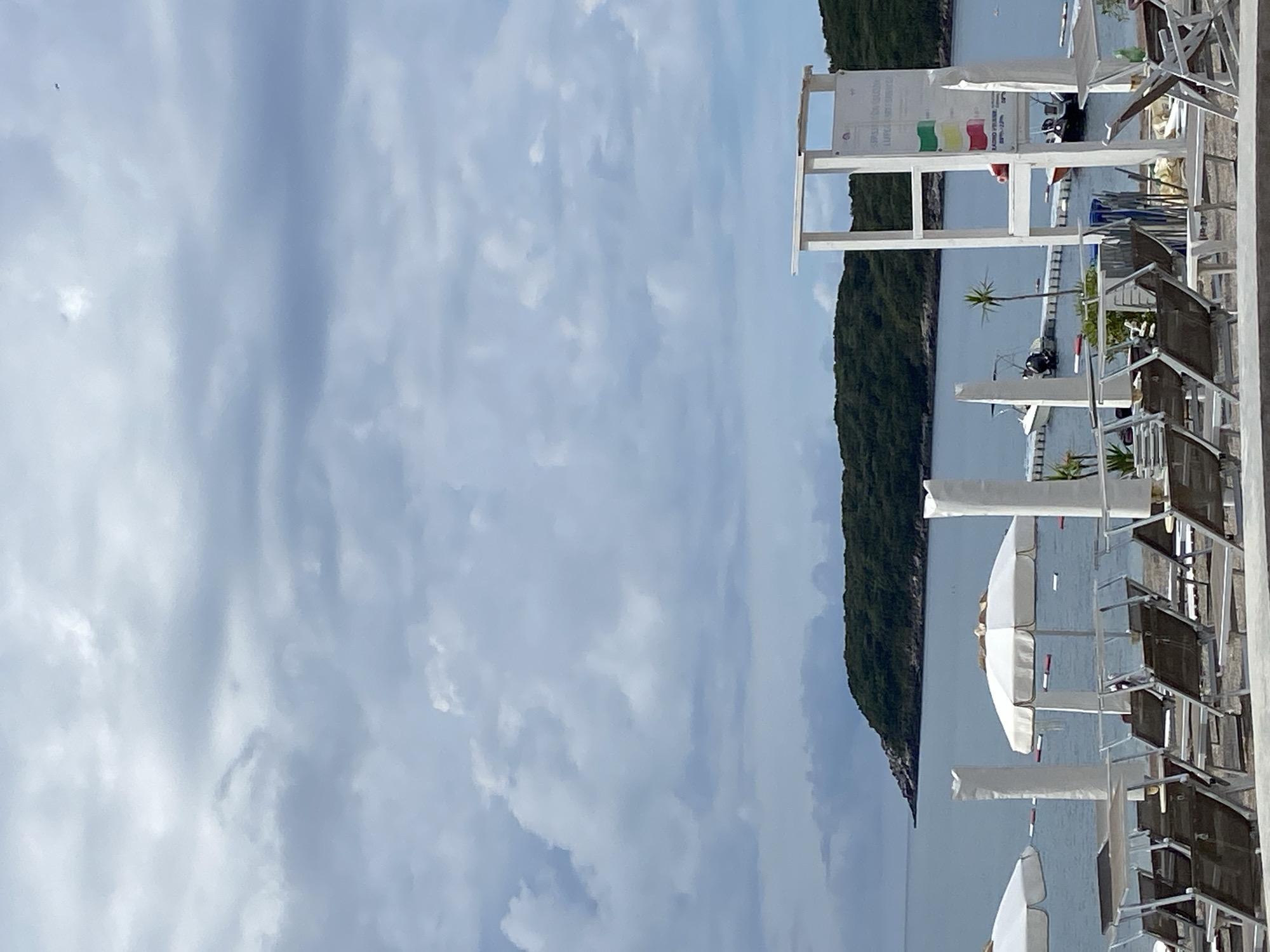 The image shows a view of the beach at Prijevor, Montenegro. The scene includes a cloudy sky over a body of water, with a tree-covered hill in the distance. Beach umbrellas and chairs are visible in the foreground, and a sign is present. Boats are present on the water.