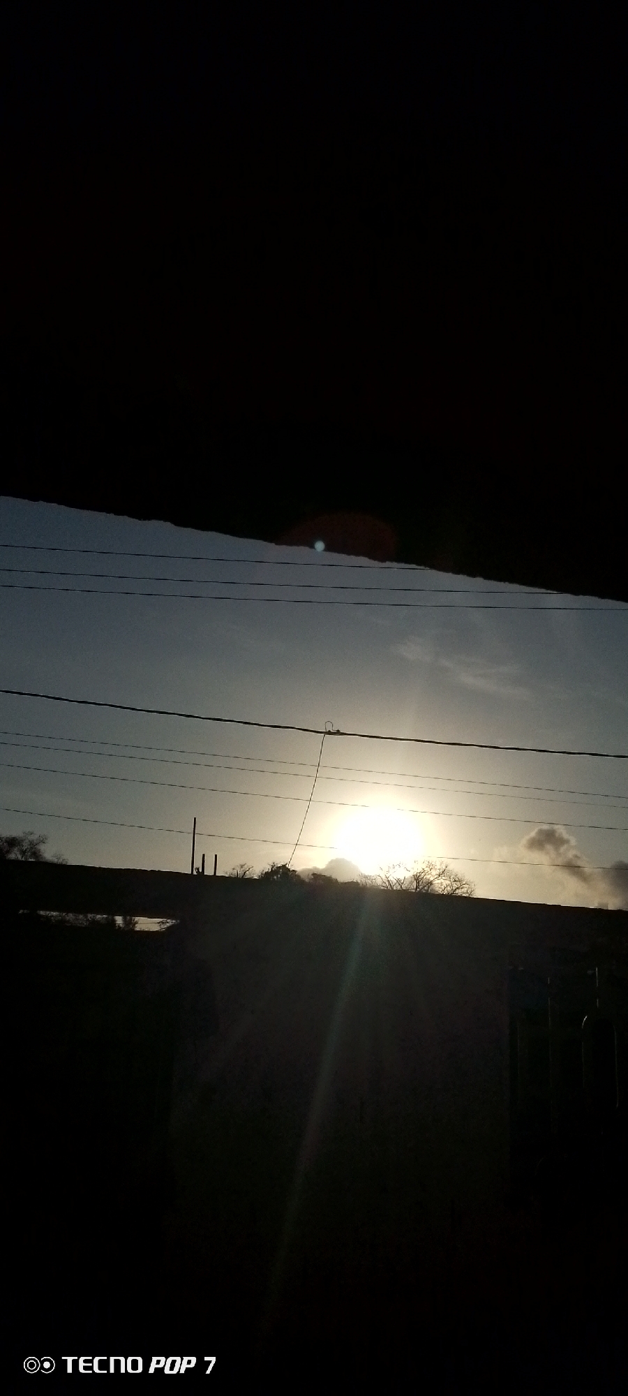 The image depicts a tranquil scene at either sunrise or sunset, with the sun positioned low on the horizon, radiating a bright, white light. The sky above is a muted blue-grey, with some lighter clouds visible, particularly around the sun. Several dark power lines stretch horizontally across the mid-ground of the frame, traversing the sky. One of these lines features a noticeable loop of cable hanging from it.
In the foreground, the top section of the image is dominated by a large, dark, irregular silhouette, likely an architectural element such as a roof or an overhang, partially obscuring the sky. Below the power lines, the landscape is presented in deep silhouette, showing what appears to be a hilly terrain or distant structures topped with dark, sparse foliage or trees. A distinct, bright blue circular lens flare is visible in the upper-left portion of the sky.
The time of day is indicated by the low position and intense glow of the sun, creating a strong contrast with the silhouetted elements. The scene conveys a sense of peaceful natural beauty. In the bottom left corner of the image, the watermark "TECNO POP 7" is visible. The location context provided is Altagracia, Venezuela.