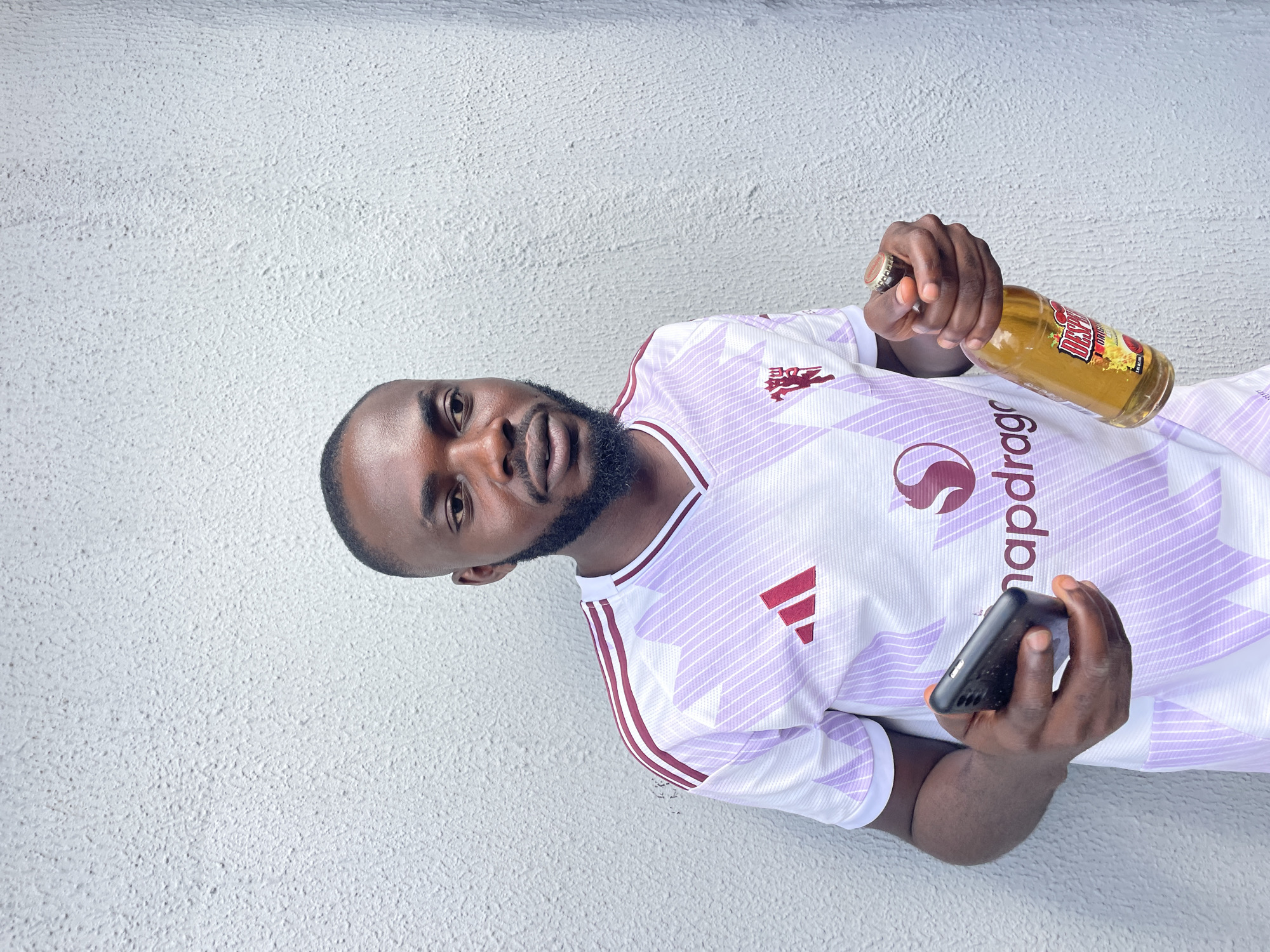 A Black male stands leaning against a textured white wall, holding a smartphone in his left hand and a bottle of Desperados Original beer in his right. He wears a white and light purple Adidas jersey with maroon accents, featuring the Adidas logo and a red Manchester United crest. The man has a beard and is looking slightly off-camera. The setting appears to be outdoors, against a plain wall that could be found in an urban environment like Jos, Nigeria.