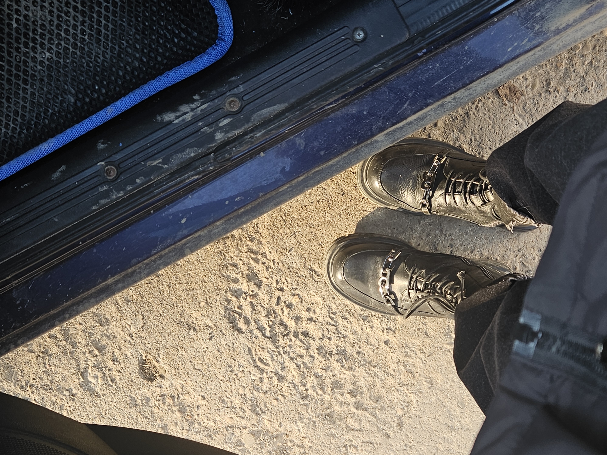 A high-angle shot depicts a person standing on a light-colored, textured surface, possibly concrete or packed dirt, next to the blue step of a vehicle. The person is wearing black boots with decorative chains and dark-colored pants. The vehicle step has a black rubber mat with a blue border. The location is unknown, and the presence of Chaikovsky, Russia, cannot be verified.