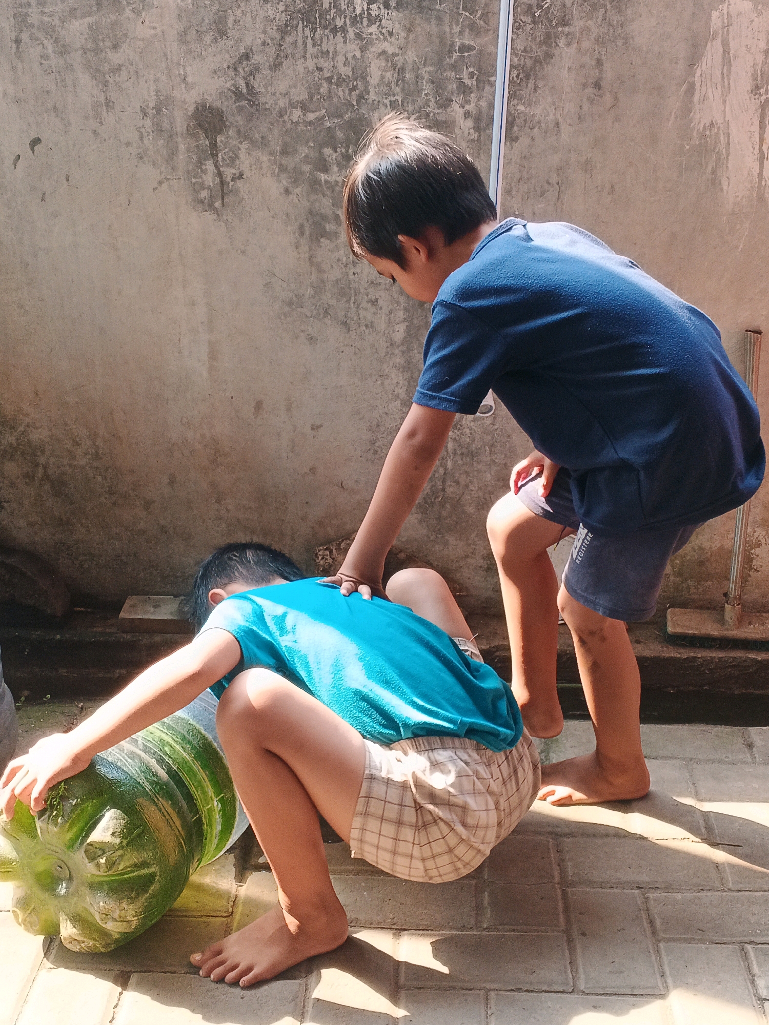 Two young boys are interacting with a large, green, ribbed squash on a paved area outdoors. The boy on the left, wearing a teal shirt and plaid shorts, is crouched down, reaching out to touch the squash with his left hand. His feet are bare. The boy on the right, wearing a dark blue t-shirt and dark shorts, is standing and leaning forward, with his right hand resting on the shoulder of the boy on the left. They are positioned against a textured, weathered concrete wall, with sunlight casting shadows on the ground. The environment appears to be a backyard or outdoor workspace in Kadu, Indonesia.