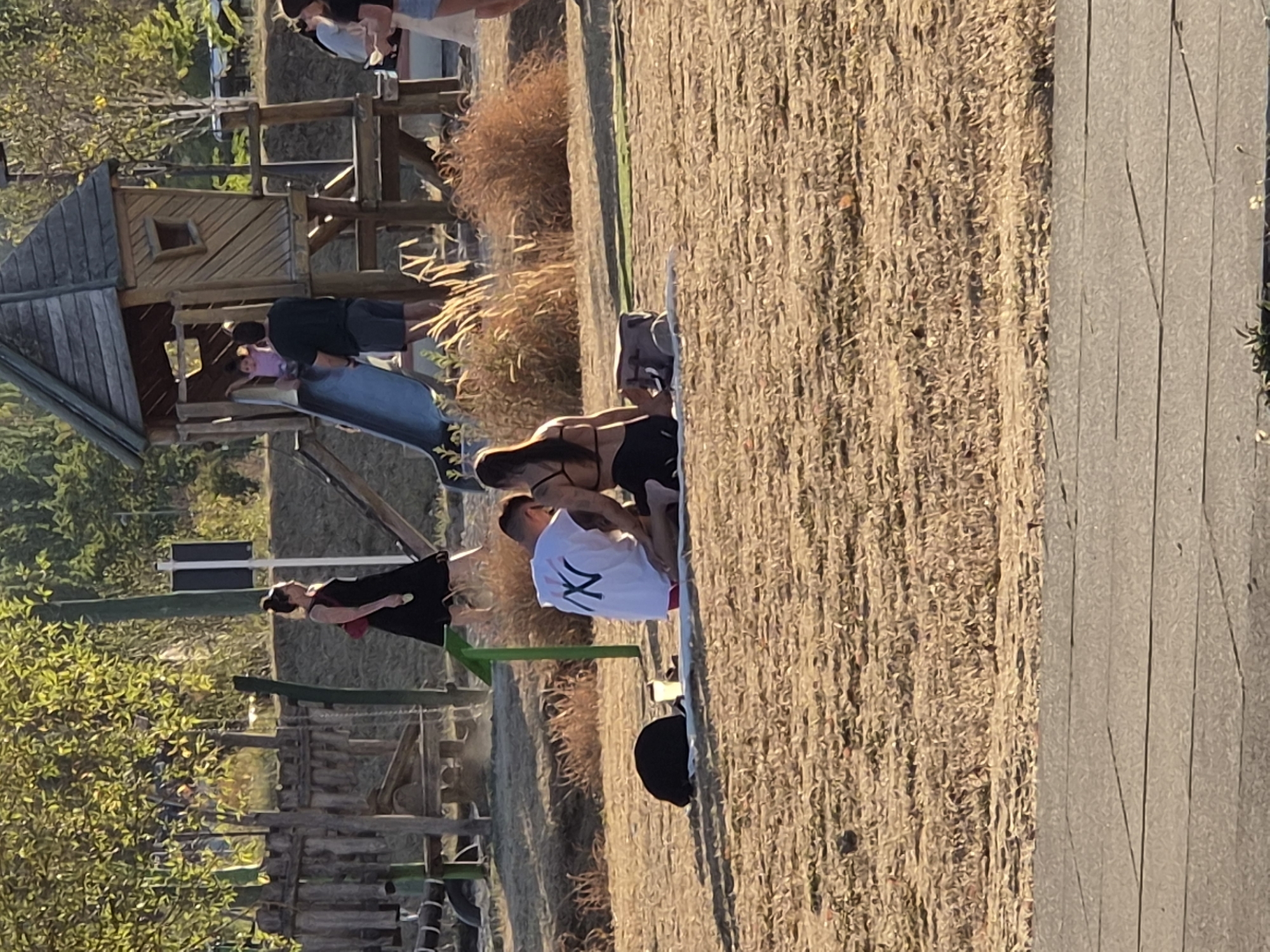 In Sanem, Luxembourg, a sunny afternoon unfolds at a park. A couple, a man and a woman, are seated on a blanket in the dry, sun-drenched grass. The man wears a white t-shirt with a black "NY" logo, and the woman sports a black bikini top and bottoms. They appear to be engaged in conversation or enjoying a moment together. Nearby, a black backpack and a drink container rest on the blanket. In the background, a wooden playground structure features a slide, with a child visible on it and an adult standing nearby. Other park-goers are scattered throughout the scene, with one woman in a black dress walking away from the camera. The environment suggests a recreational area with trees and dry vegetation.
