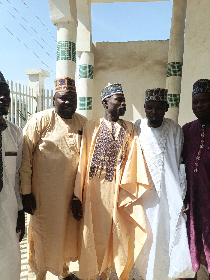Five adult males are observed standing outdoors in the city of Gawuna, Nigeria. They are arranged in a line, posing under bright daylight, with shadows indicating an overhead sun source. All subjects are dressed in traditional Nigerian attire, consisting of long-sleeved robes and intricately embroidered caps.
From left to right:
1. The first male, partially cropped on the left, wears a light-colored tunic with dark trim on the chest and a dark cap.
2. The second male wears a light tan long tunic and a square, multi-colored embroidered cap, looking directly forward.
3. The third male, centrally located, wears a light tan long tunic with extensive blue and gold chest embroidery and a multi-colored cap, facing slightly to his right.
4. The fourth male wears a white long tunic with subtle white geometric embroidery on the chest and a multi-colored cap, facing slightly to his left.
5. The fifth male, partially cropped on the right, wears a deep maroon long tunic with light-colored chest embroidery and a dark cap.
The background consists of a white metal fence on the left and a beige building facade with prominent white cylindrical pillars. These pillars feature horizontal bands of green square tiles. Above the structure, a clear blue sky is visible through an open area, with overhead power lines present in the upper left quadrant. No overt interactions or specific actions are discernible beyond posing for the photograph.