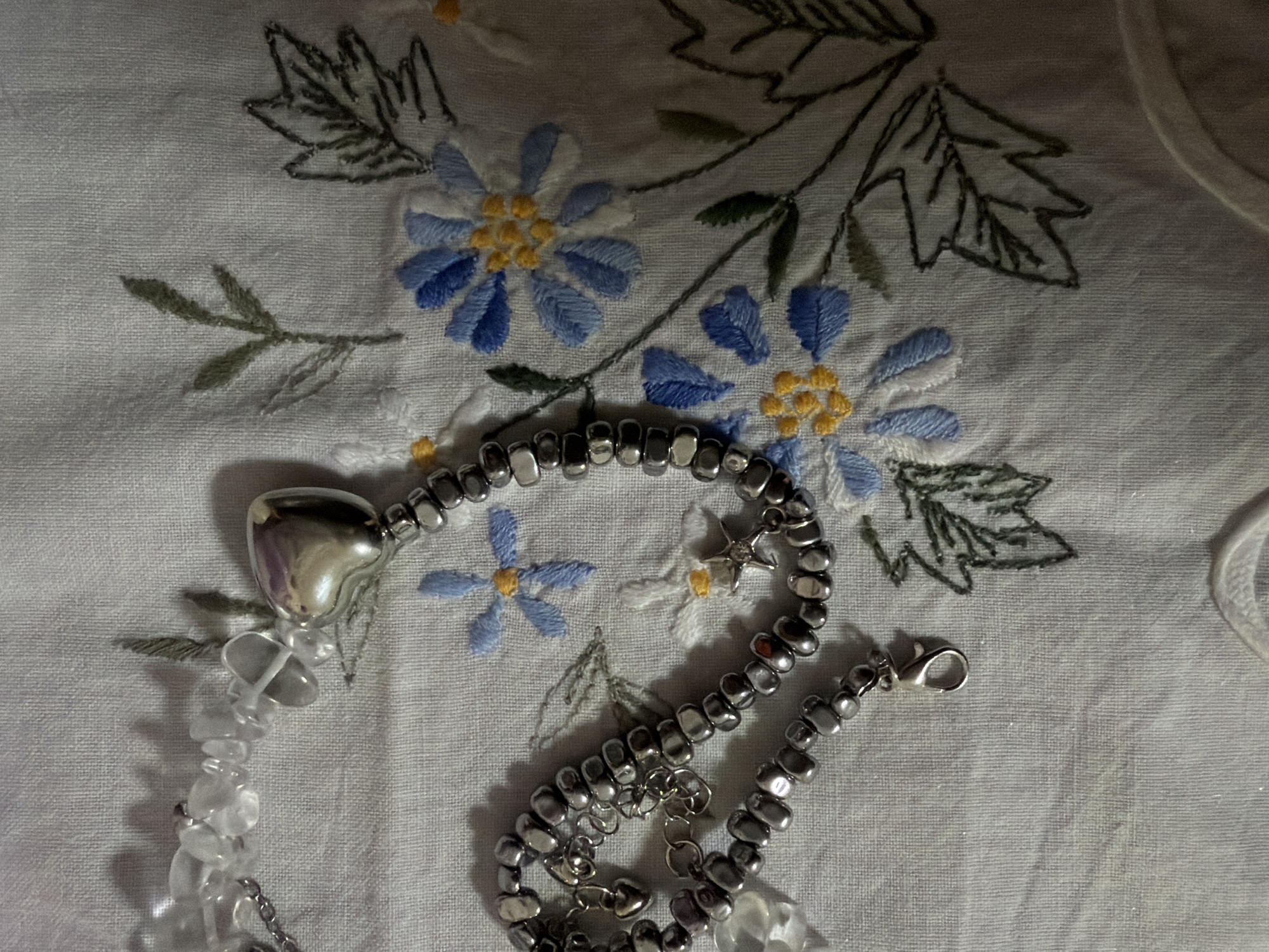 A close-up shot shows a piece of jewelry, possibly a necklace or bracelet, lying on top of a white embroidered cloth. The cloth features a pattern of blue flowers with yellow centers and green leaves. The jewelry is comprised of silver-toned beads, a heart-shaped pendant, and transparent, crystal-like beads. Some of the metal beads have small dangling charms attached.
There is no information present that would allow for the identification of any persons or their actions, nor information about the location in Izhevsk, Russia.