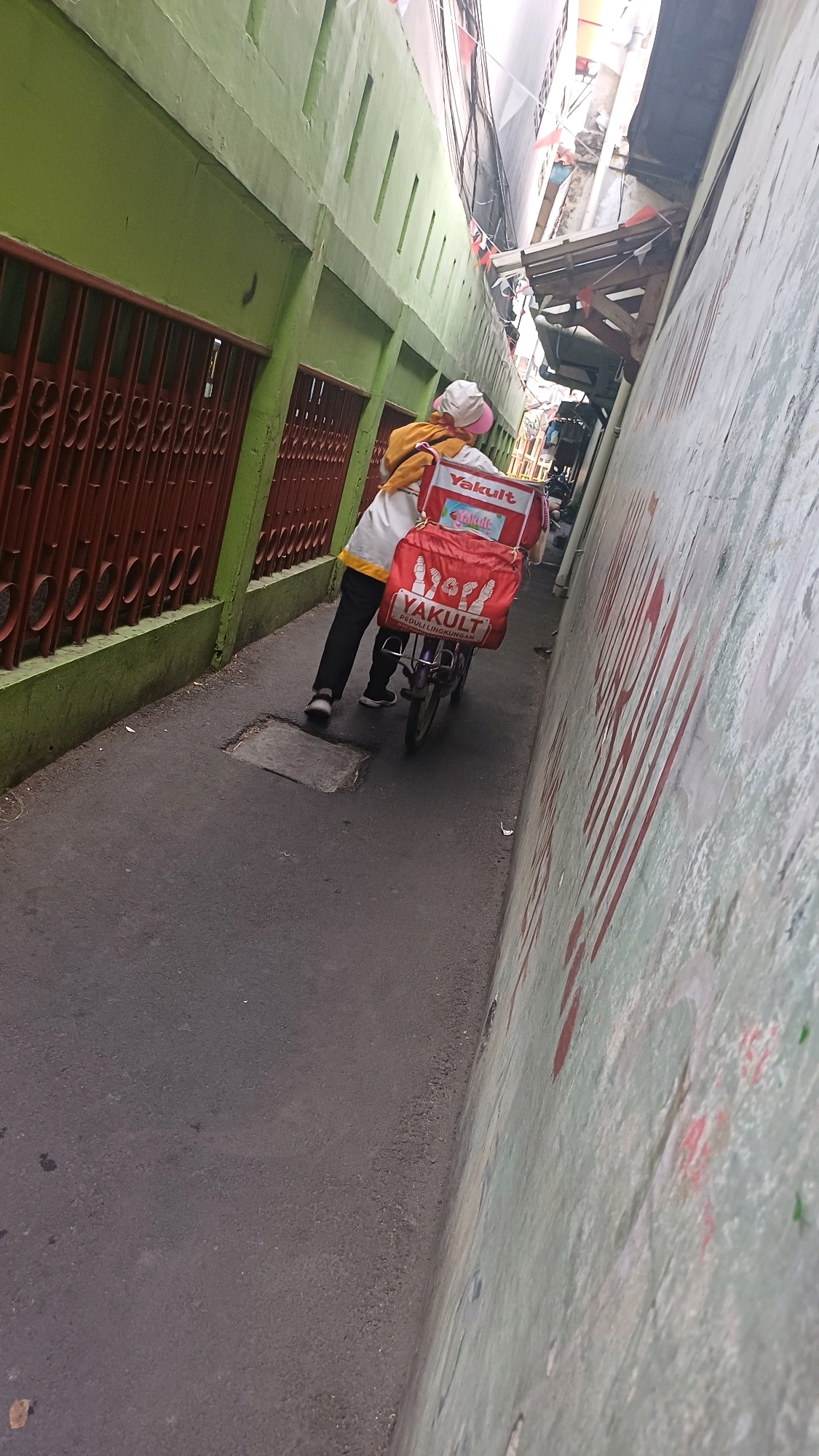 A person wearing a yellow jacket and a white hat rides a bicycle down a narrow alleyway. The bicycle has a large red insulated box on the back with "YAKULT" branding. The alleyway is flanked by buildings; the left side is painted green with a red barred fence, and the right side is a light green wall with red graffiti. The ground is asphalt. Red and white Indonesian flags are strung overhead, suggesting a festive occasion. The scene appears to be in a densely populated urban area.