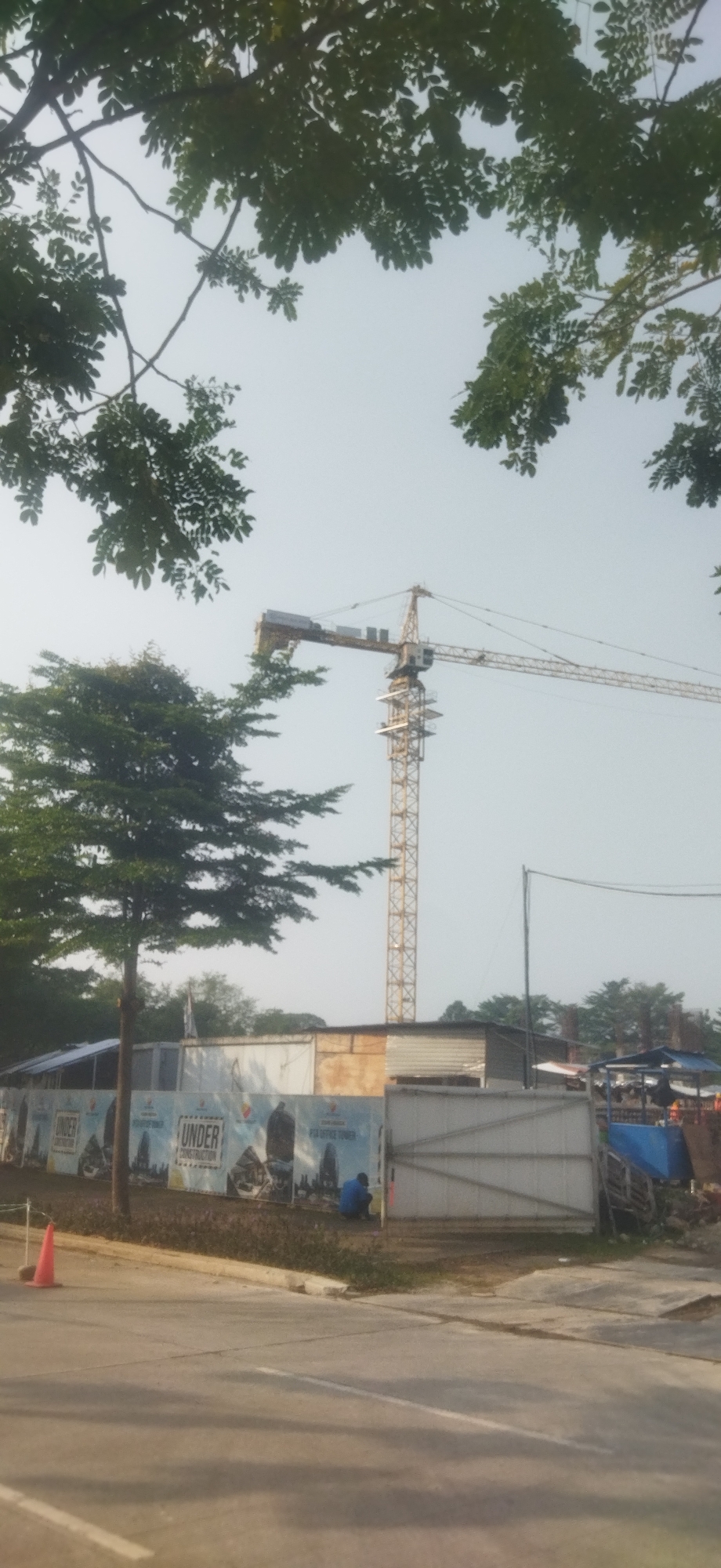 The image shows a construction site in Depok, Indonesia, dominated by a large yellow tower crane. The crane's boom extends horizontally across the frame, with its jib reaching towards the right. In the foreground, a paved road runs horizontally, with a traffic cone on the left edge of the image. Behind the road, a construction fence is visible, featuring large banners that read "UNDER CONSTRUCTION" and show images of skyscrapers. A single person, wearing a blue shirt, is seated on the ground near the fence, facing away from the camera. Several trees with green foliage frame the top and left sides of the image. The sky is overcast and a hazy light illuminates the scene. Various construction materials, including a blue tarp and wooden pallets, are present in the background.