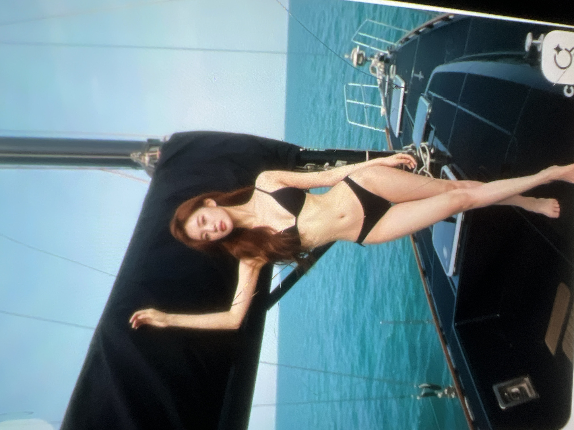 The image shows a woman wearing a black bikini standing on a sailboat. She has long, brown hair and is looking at the camera. Her left arm is raised, resting on a black sail behind her. The sailboat is on a body of water with a clear blue sky above. The water appears to be calm and turquoise. The deck of the sailboat is black and has metal railings. The image is taken from a slightly low angle, emphasizing the woman and the sailboat against the backdrop of the sky and sea. The location in the city of Kara-Dıykan, Kyrgyzstan is not evident from the image.