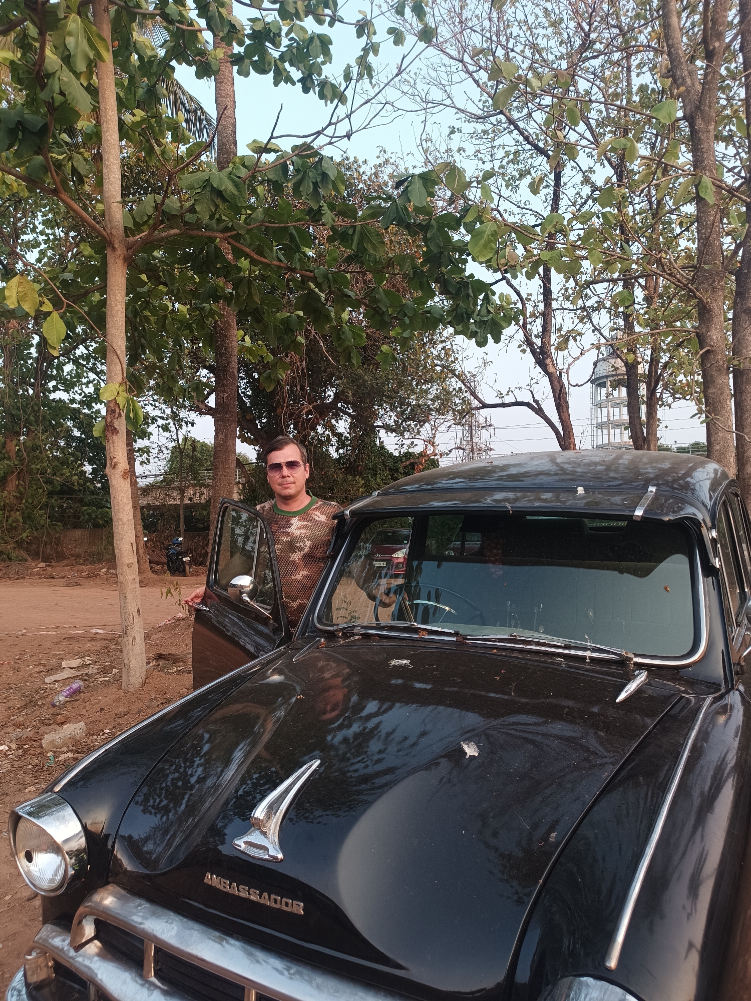 A medium outdoor shot features a man in sunglasses standing next to a vintage black Ambassador car. He is opening the driver-side door of the car. The car's front grill clearly displays the word "Ambassador".
The scene is set in a partially unpaved area. Behind the man and car, there are trees and glimpses of buildings and infrastructure, including a water tower. A motorcycle is parked in the background. The city location is identified as Kalamassery, India.