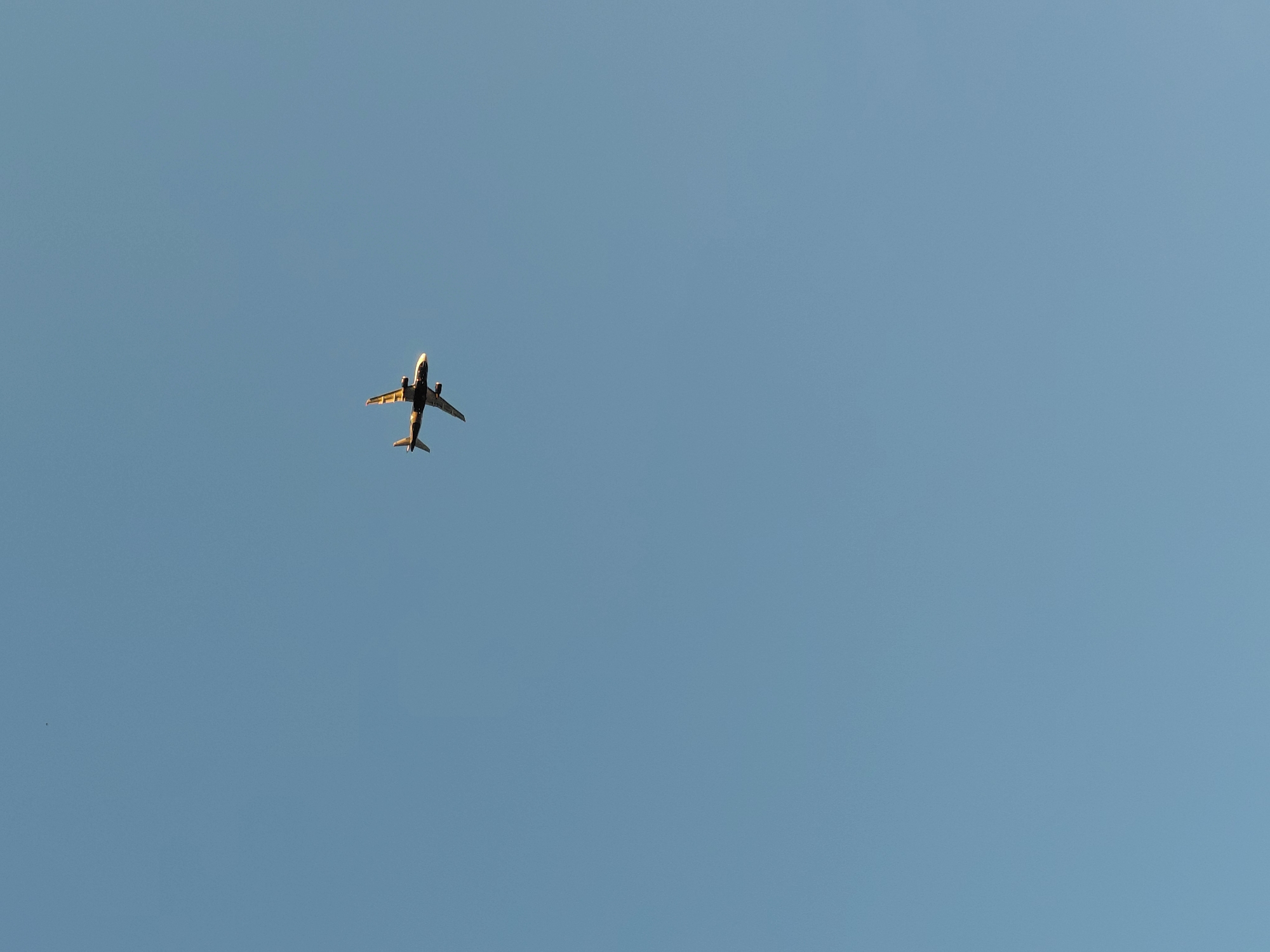 An airplane is seen flying in a clear blue sky. The airplane is a commercial jet with two engines mounted under its wings. It is angled slightly upwards and to the left, appearing to be ascending. The sun is casting a warm glow on the underside of the aircraft, suggesting it is either early morning or late afternoon. The sky is a uniform, soft blue, indicating fair weather with no clouds visible. The scene is devoid of any other objects or elements, focusing solely on the aircraft against the vast expanse of the sky. The perspective is from below the plane, looking up. There is no visible text in the image. The image captures a moment of flight, evoking a sense of travel or transition.