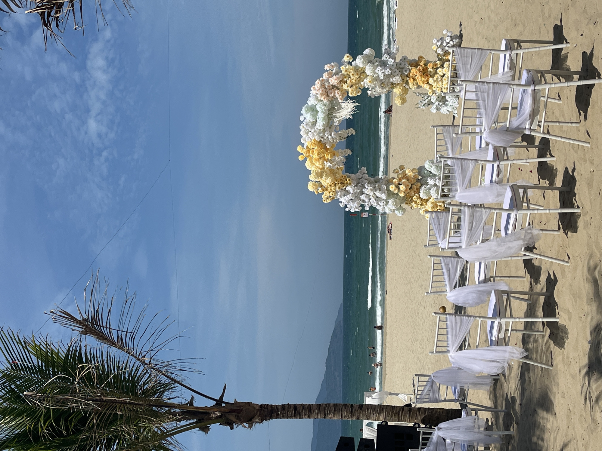 This image depicts a serene beach wedding or ceremonial setup on a bright, sunny day in Đà Nẵng, Vietnam. The scene is dominated by a large, circular floral arch on the sandy beach, adorned with an abundance of white, yellow, and light pastel flowers, facing the ocean.
In front of the arch, neat rows of white chairs, elegantly draped with flowing white tulle fabric, are arranged on the sand, suggesting seating for guests. An aisle separates the two sections of chairs, leading towards the floral centerpiece and the sea. The bright sunlight casts distinct shadows from the chairs onto the sand.
To the left of the setup, a tall palm tree with a mix of green and dried fronds stands prominently, its canopy partially framing the sky. Thin wires are visible stretching across the clear blue sky, which features a few scattered clouds. A small helicopter can be faintly discerned in the distance, high in the sky.
In the background, the turquoise waters of the ocean gently lap the shore, with several individuals visible swimming or wading near the water's edge. Distant mountains rise from the horizon, providing a scenic backdrop to the coastal setting. Dark equipment, possibly speakers or sound systems, are partially visible near the base of the palm tree. The time of day appears to be mid-morning or early afternoon, indicated by the strong, direct sunlight and clear conditions, ideal for an outdoor event. No visible text is present in the image.