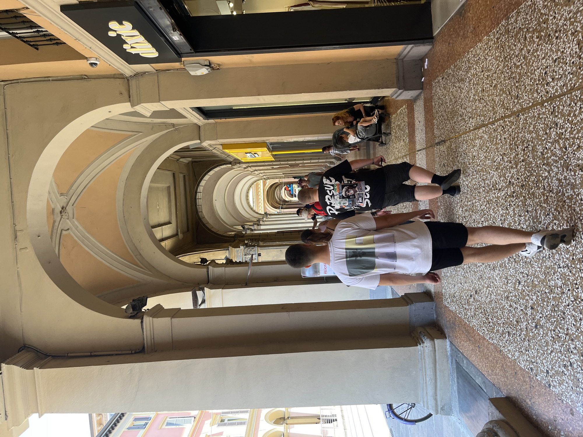 The image shows a long, covered walkway in Bologna, Italy, characterized by repeating arches that create a tunnel-like perspective. The walkway is paved with a speckled pattern of white and brown stones. A few people are walking along the walkway. Two young men are walking in the foreground. One is wearing a white t-shirt and black shorts; the other is wearing a black t-shirt and black shorts. Further down the walkway, a person pushes a stroller. Stores line one side of the walkway with signs visible. One sign reads "Dixie." A security camera is visible above one of the arches. The walkway is lit by natural light filtering through the arches and openings. The image is taken at ground level, looking down the length of the walkway.