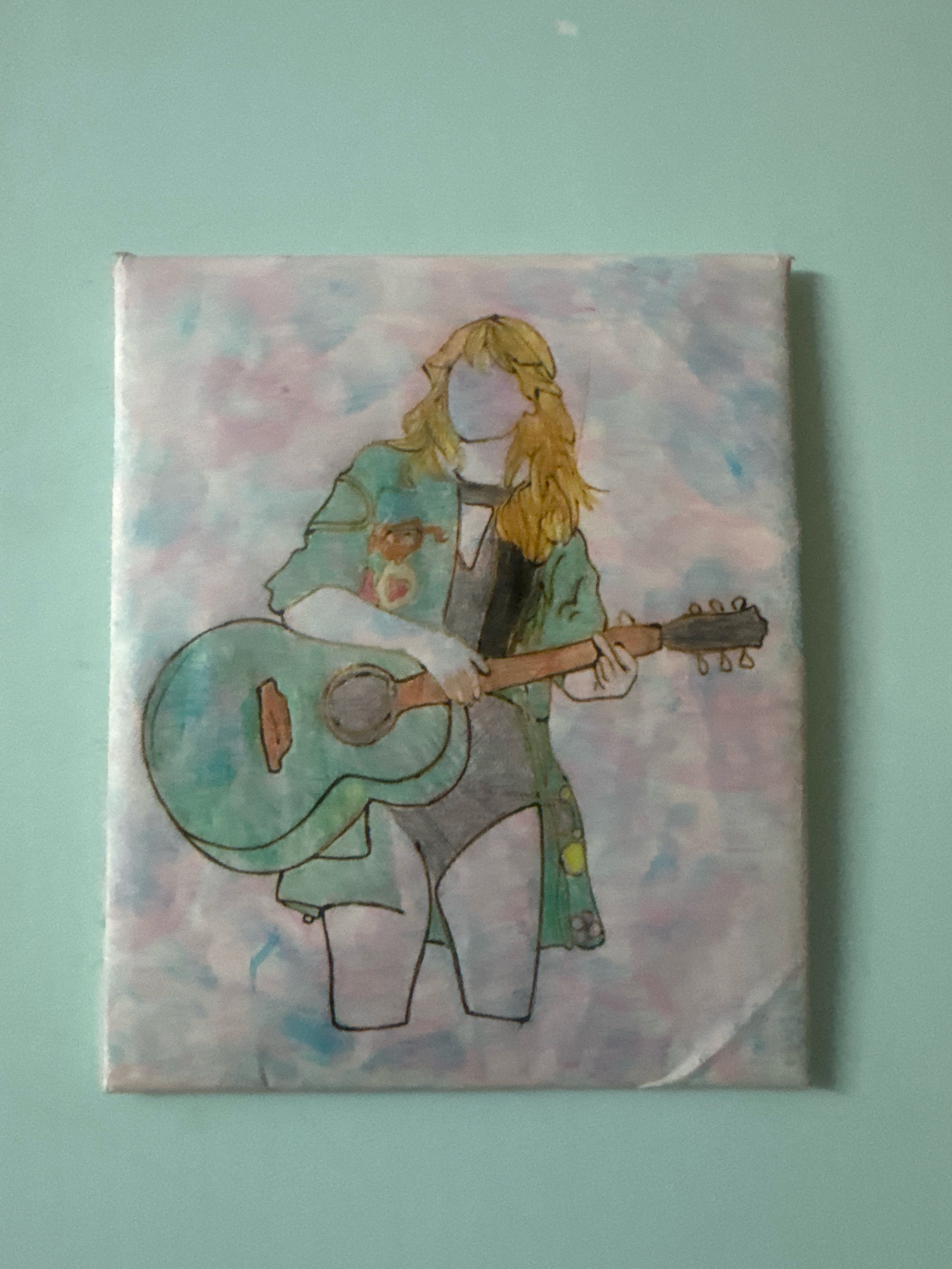 A painting of a person playing a guitar is hung on a pale blue wall. The painting is on a canvas and features a stylized figure with blonde hair, wearing a green jacket over a dark outfit, and holding a light blue acoustic guitar. The background of the painting is a wash of light pink and blue colors. The bottom right corner of the canvas appears to be slightly peeling away from the frame. The painting is centrally located on the wall. There is no identifiable information about the location in Nagamalli Thota, India.