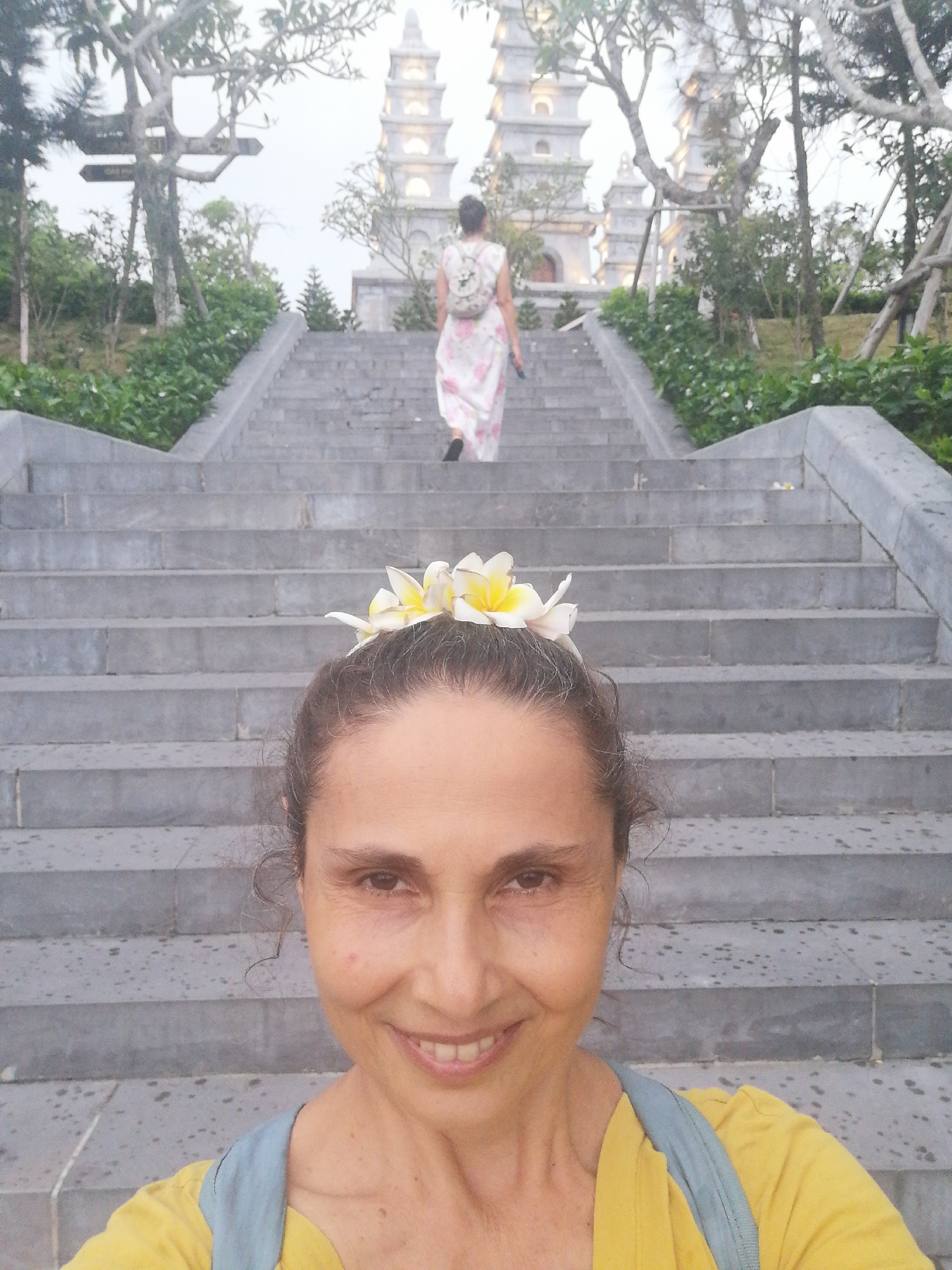 A selfie shows a middle-aged woman with a crown of frangipani flowers in her hair standing at the bottom of a long, wide, stone staircase leading to a religious structure in the background. The woman has dark hair and light brown skin. She is wearing a yellow shirt with a light blue strap over her shoulder. She is smiling at the camera. The staircase extends upwards and appears to lead towards a temple-like structure with multiple tiered pagodas. The steps are grey and show signs of being damp or wet. In the background, a woman in a pink and white patterned dress and a backpack is ascending the stairs. The stairs are lined with greenery. Trees frame the sky above. The photograph was taken in Hong Gai Ward, Vietnam.