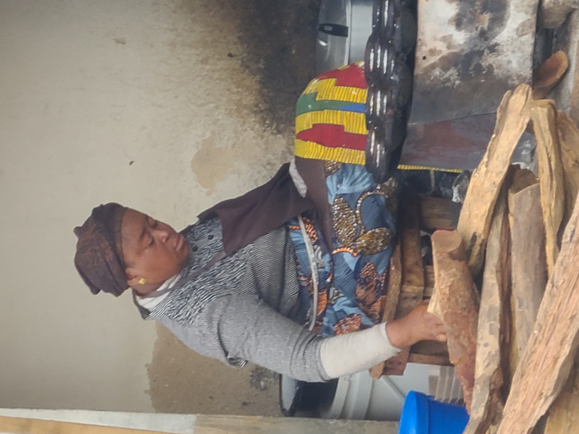 The image depicts a woman seated on a stool in front of a cooking area. She is wearing a headscarf, a striped shirt, and a patterned skirt. Her hands are resting on her lap. In front of her is a traditional stove with cooking pots. In the foreground, there are pieces of firewood. The background appears to be an outdoor setting with a textured wall and a large white bucket to the left. This scene is characteristic of a street food vendor in Jos, Nigeria, where such cooking setups are common.