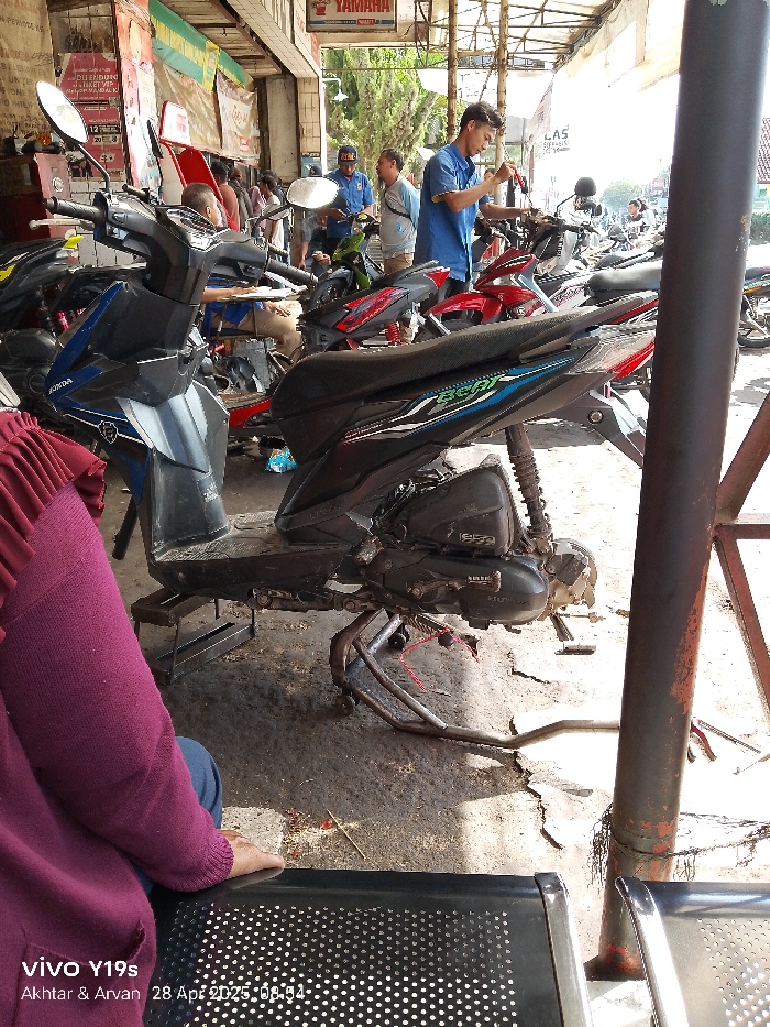 The image shows a motorcycle parked on the side of a street. The motorcycle is a Honda Beat and is black in color. The motorcycle is on a metal kickstand and its engine is exposed. The motorcycle is parked next to a metal pole that has a sign that says "YAMAHA" on it. There are other motorcycles parked in the background. The scene is located in Kecamatan Haurgeulis, Indonesia, based on coordinates (latitude: -6.45, longitude: 107.942). A person in a maroon sweater is seated on a black bench in the foreground. The person's hand is resting on the bench. The pavement is dirty and there is a shadow on the ground cast by the motorcycle and the bench. There are multiple objects lying on the ground, including a motorcycle kickstand, a rope, and a small piece of debris. There are multiple people in the background, including a man in a blue shirt working on a motorcycle. The background is blurry due to the distance from the viewer.