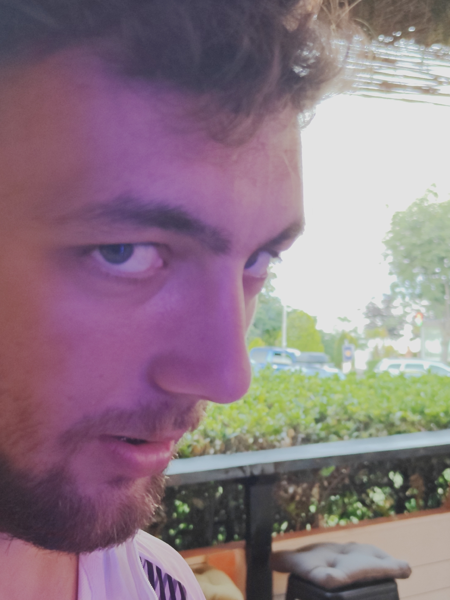 The image is a close-up, low-angle shot of a young man's face, with a portion of his body and clothing visible. He appears to be outdoors, with blurred greenery and a hint of buildings and vehicles in the background. The lighting suggests either twilight or an artificial purple light source. He has curly brown hair, a light beard, and is looking towards the right side of the frame. His left eye is clearly visible, and his mouth is slightly open. He is wearing a white jacket with black and white striped detailing on the collar. The overall impression is a casual, candid shot. It is not possible to determine the specific location within the city of Tsarevo, Bulgaria, from the visual information provided in the image.