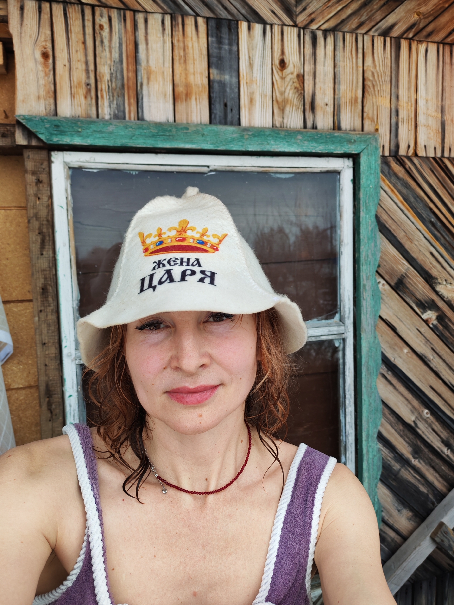 A woman is photographed outdoors, seemingly near or within a wooden structure. She wears a white felt hat with a crown image and Cyrillic text that translates to "Queen of the Tsar." She also wears a purple top with white trim and a red necklace. Her hair is wet. The background consists of a wooden wall and a window with a green frame. The location is Chaikovsky, Russia.